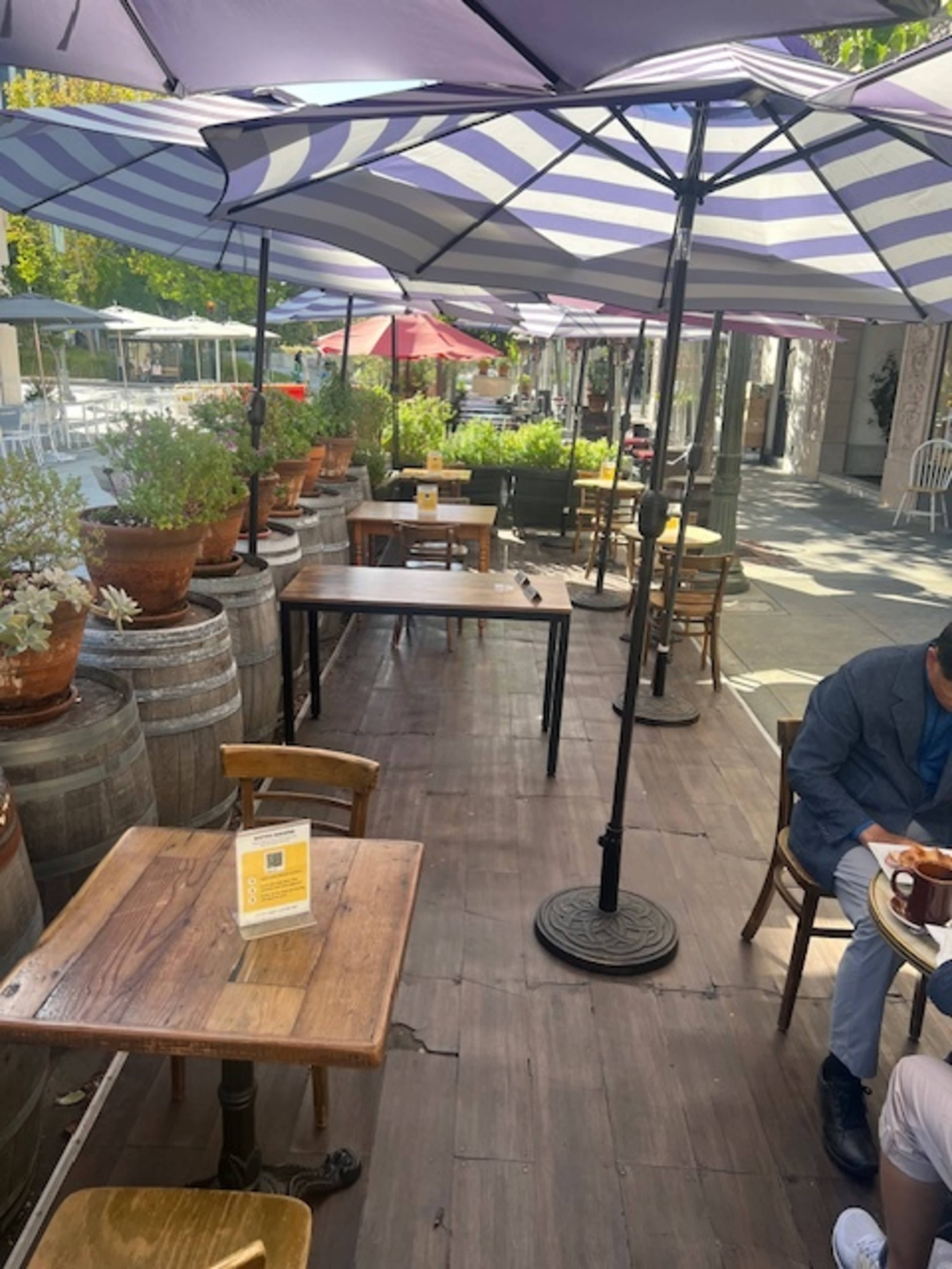 The image shows an outdoor seating area at a café with wooden tables, striped umbrellas, and potted plants.