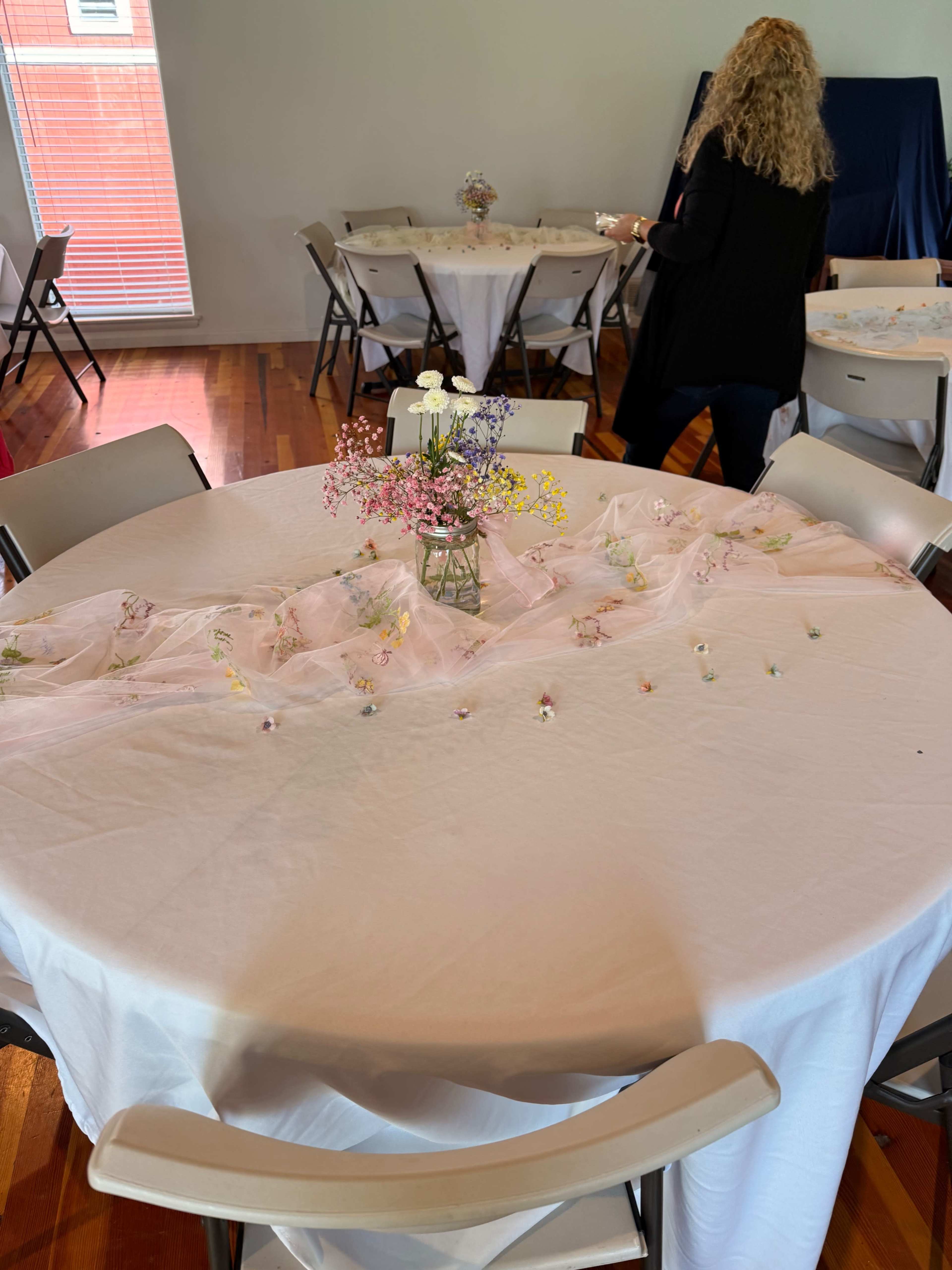 A circular table with a white tablecloth is decorated with small flowers and a glass vase, while a person arranges items at a nearby table in a bright room.