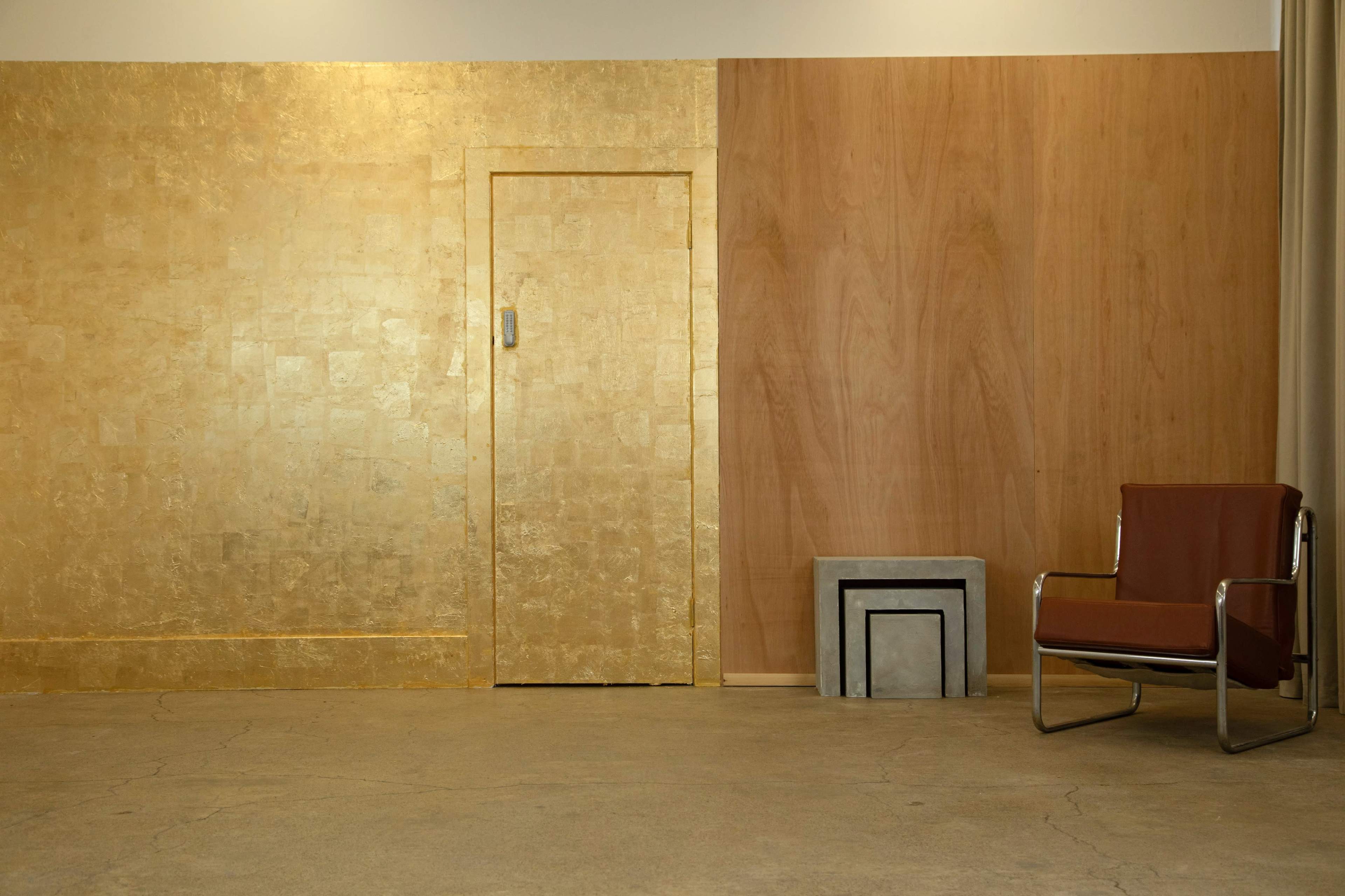 A minimalist interior with a gold-leaf wall on one side, a wooden wall on the other, a small concrete table, and a brown leather chair.