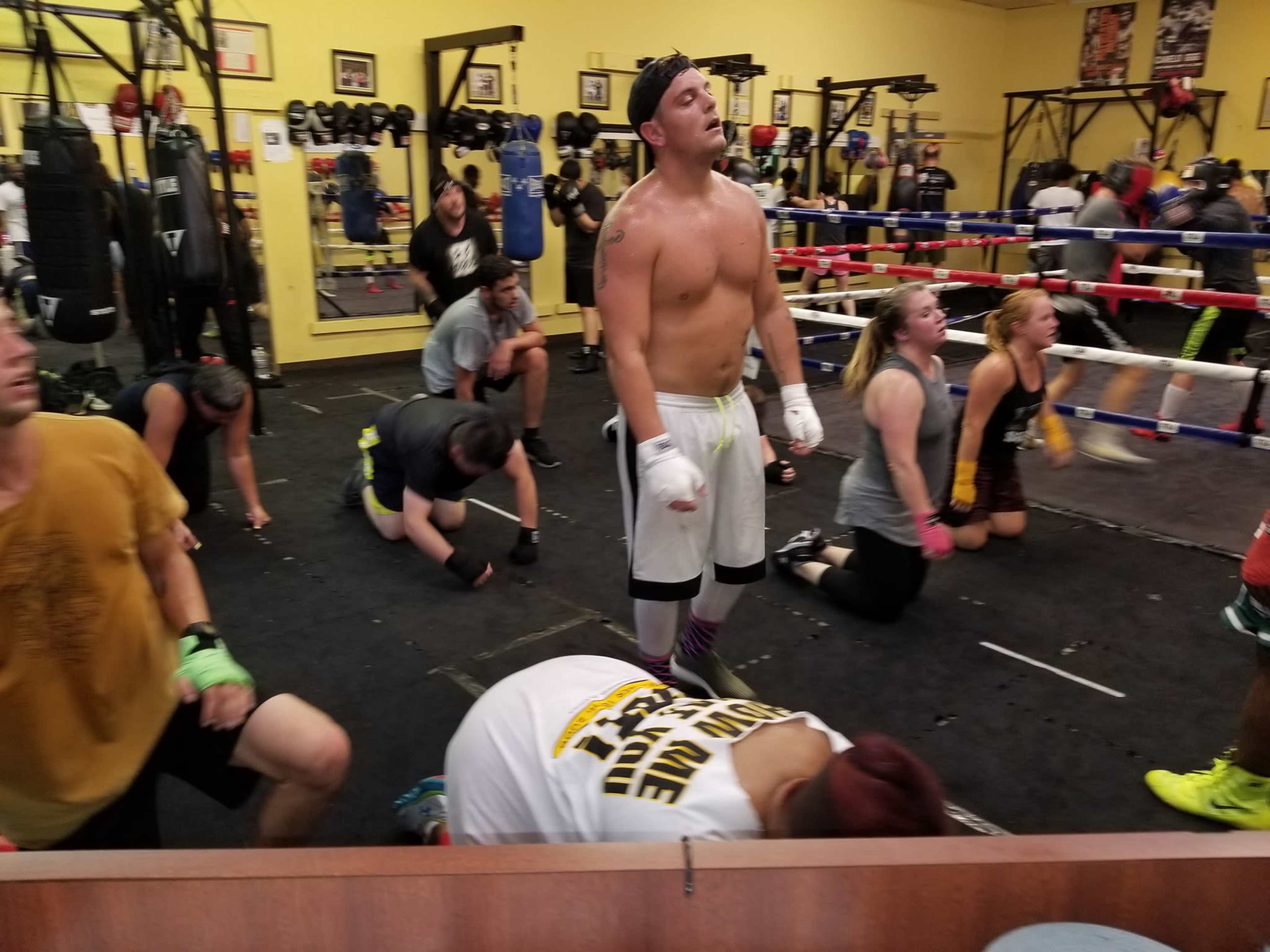 A group of individuals is training in a boxing gym, with some participants on their knees and others standing, in a lively practice session.