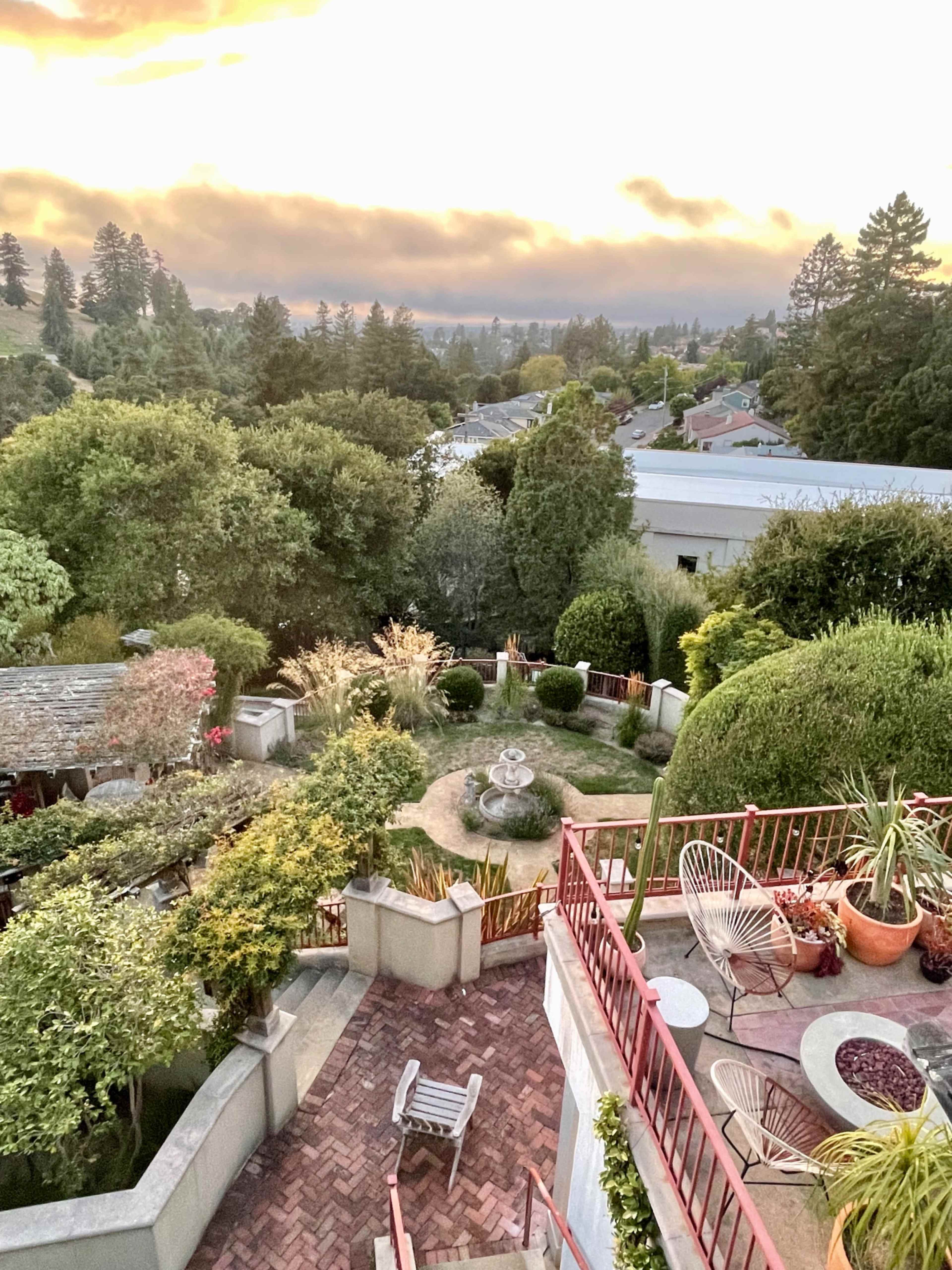 The image shows a landscaped garden with pathways, various plants, and a fountain, set against a backdrop of trees and a residential area under a sunset sky.