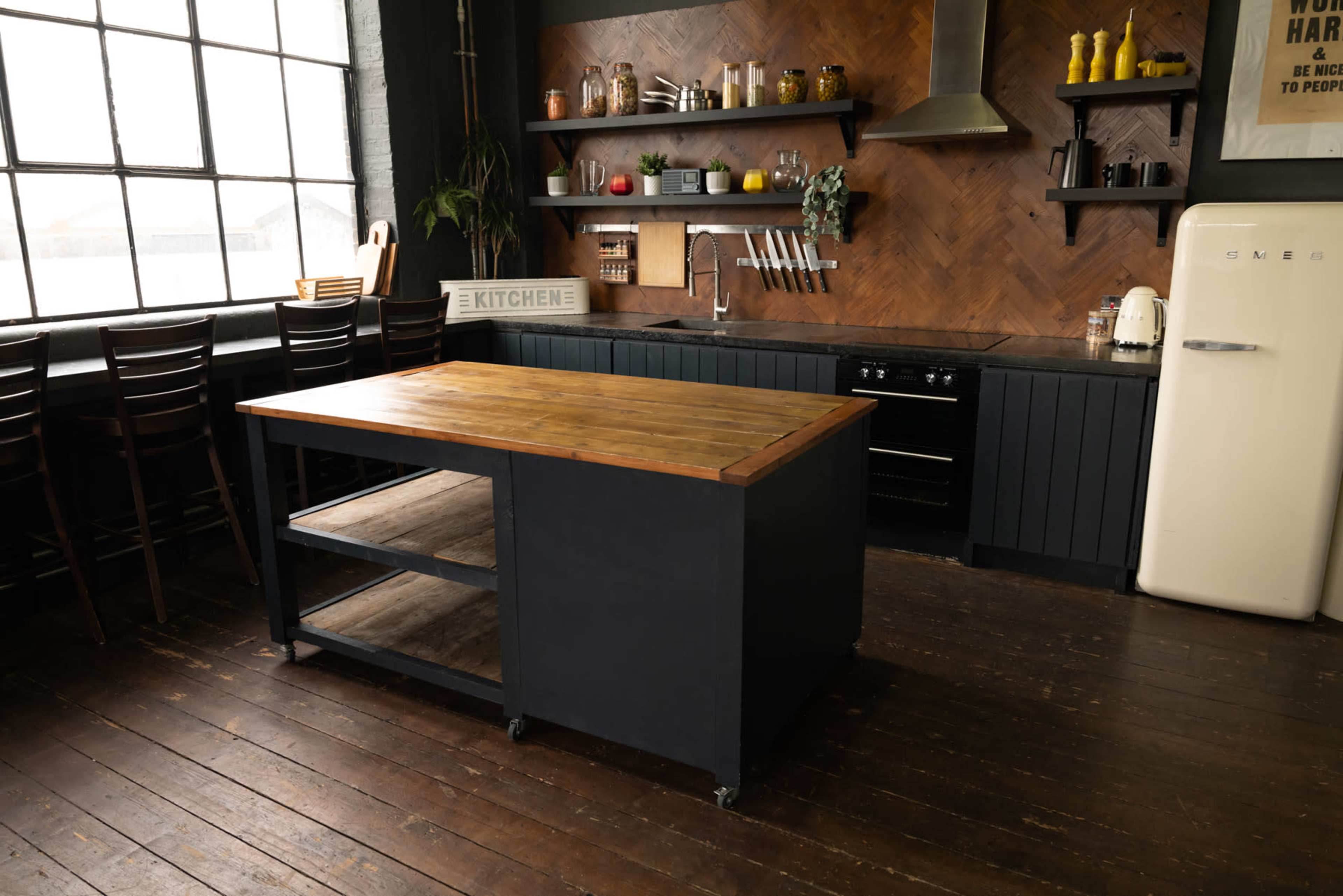 A modern kitchen featuring a wooden island with a dark base, black cabinets, and a vintage-style refrigerator, all set against a backdrop of a large window and wooden flooring.