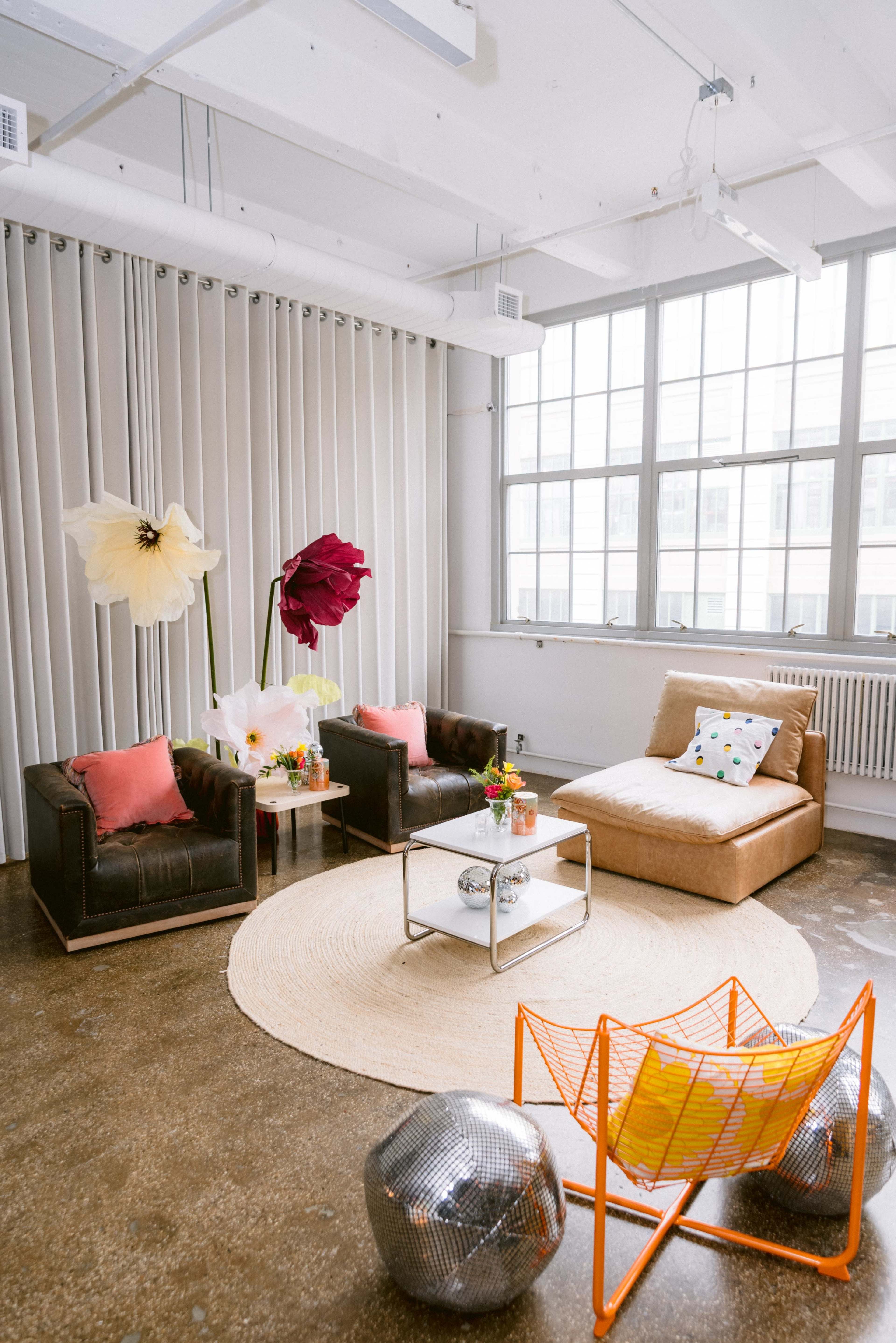The image shows a modern living space featuring two brown leather armchairs, a beige lounge chair, a round rug, a coffee table, large fabric flower decorations, and colorful cushions.