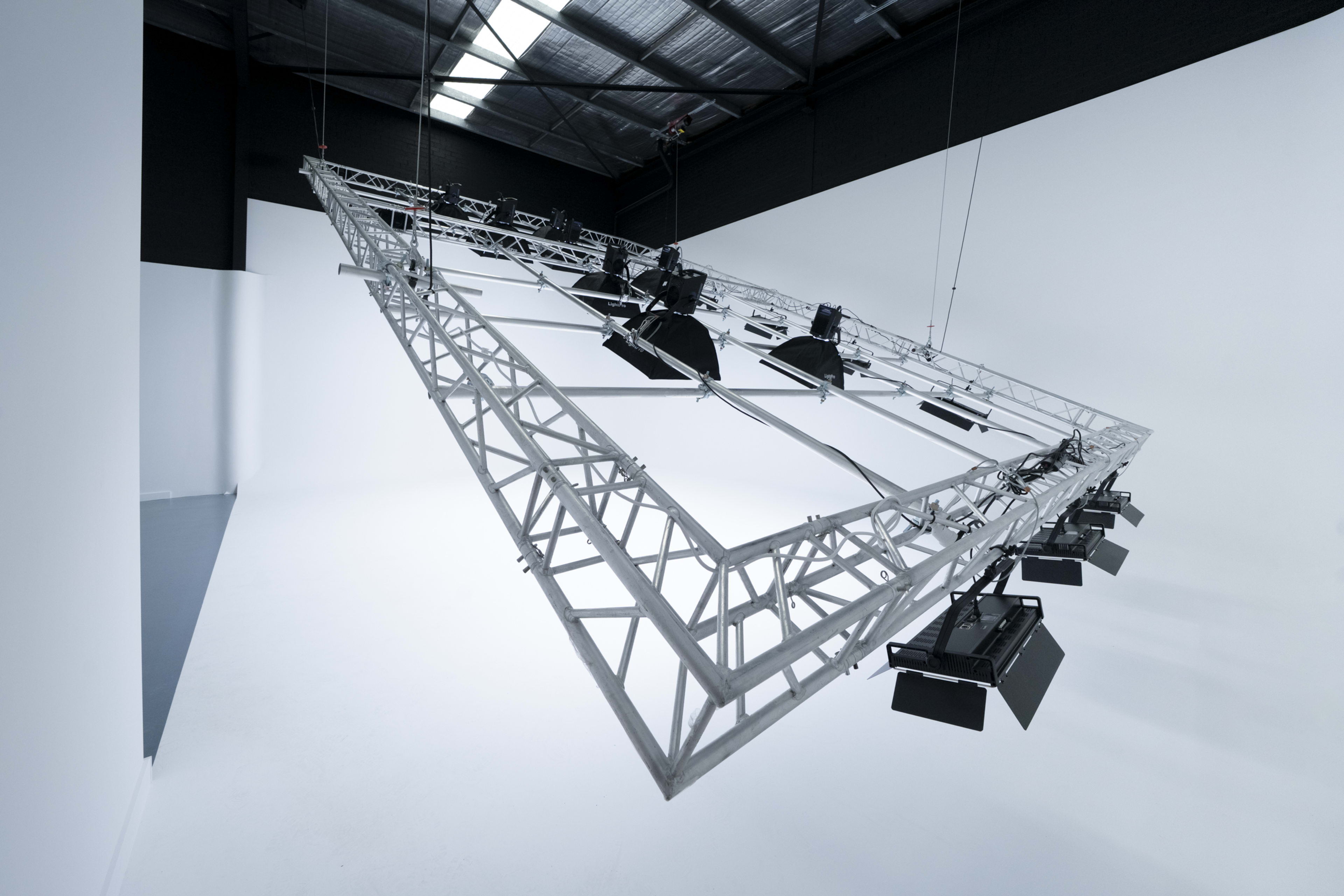 A large, angled metal truss structure supports multiple stage lights in an empty warehouse-like space.