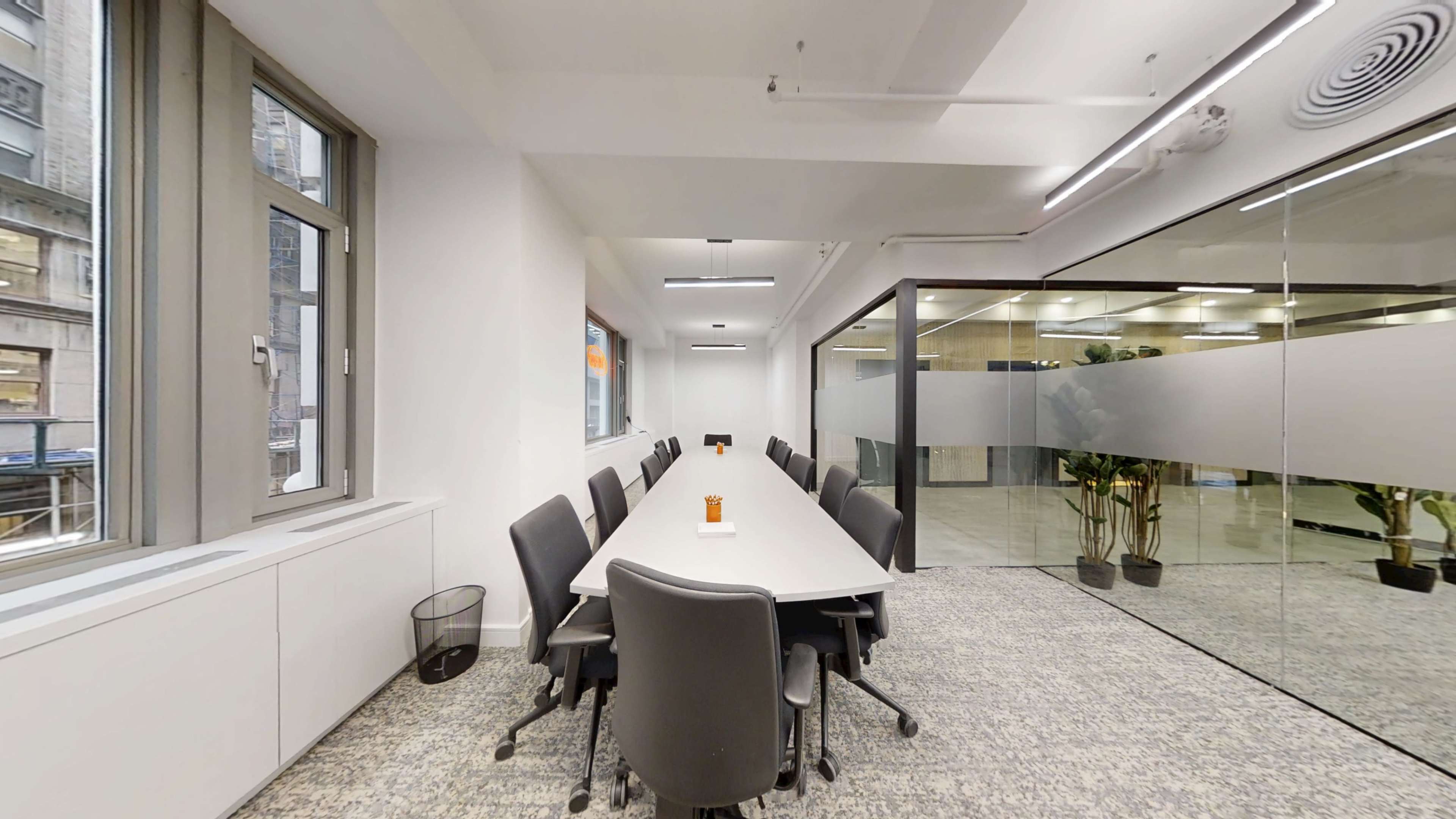 A long modern conference table is set with chairs around it, situated in a well-lit room with large windows and a glass partitioned area.