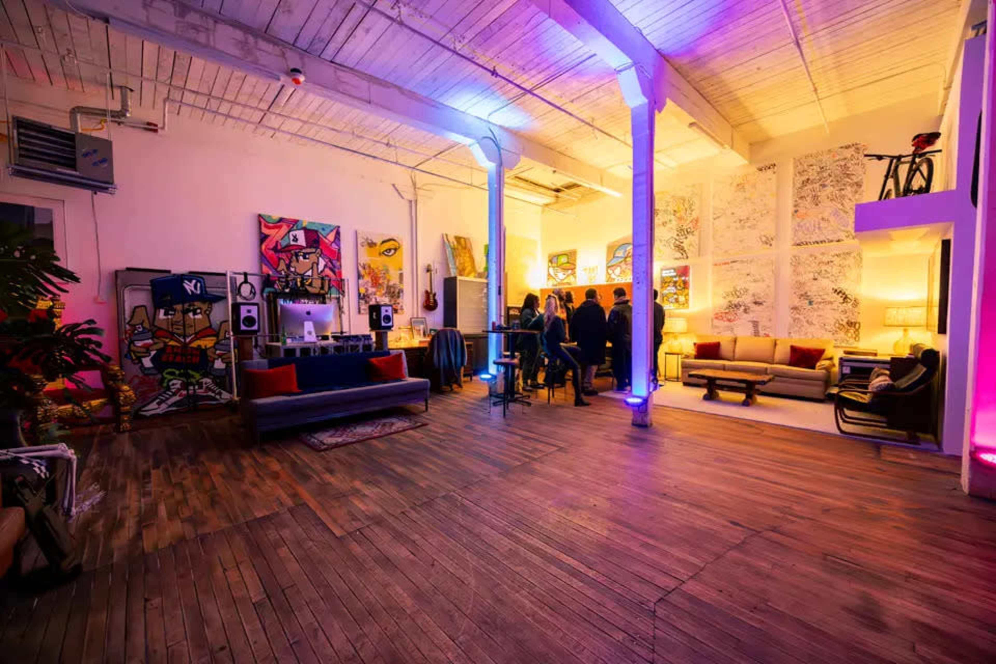 The image shows a spacious, well-lit room with modern furniture, vibrant wall art, and groups of people engaged in conversation.