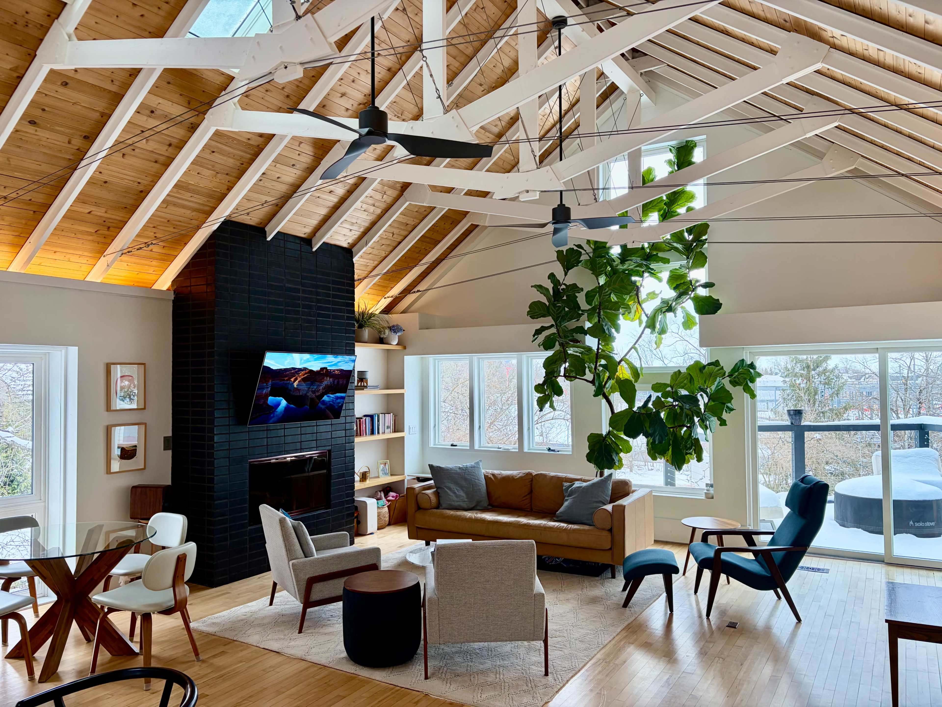 The image shows a modern living room with a high, wooden-beam ceiling, large windows, a black fireplace, and a mix of contemporary furniture including a couch, chairs, and a coffee table.