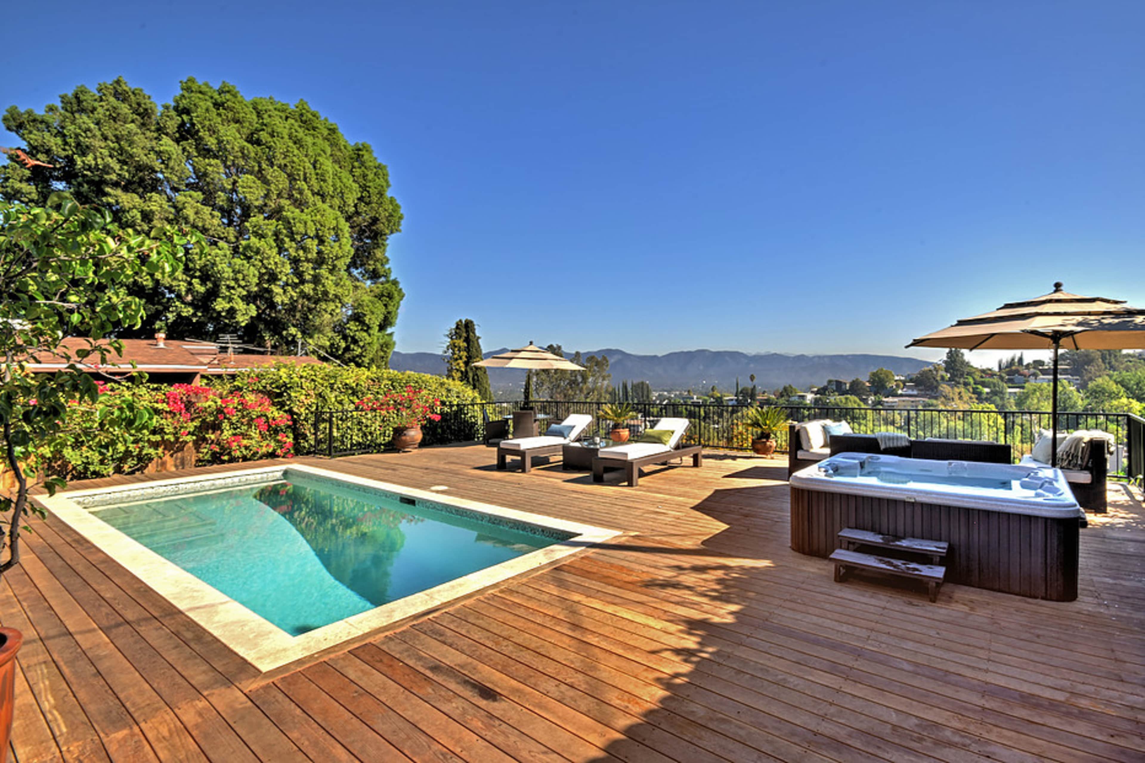 The image shows a wooden deck with a swimming pool and a hot tub surrounded by greenery and mountains in the background.