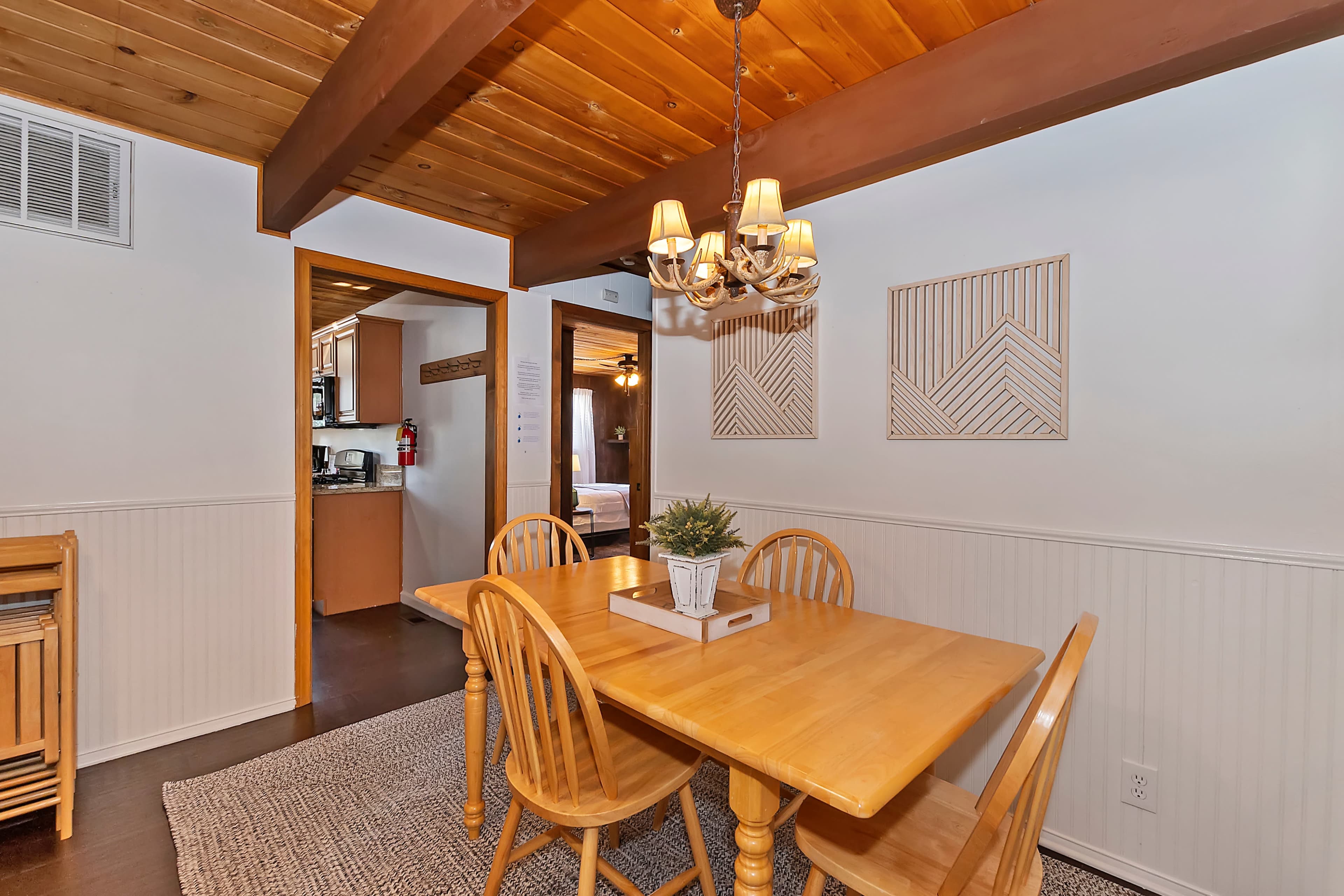 A wooden dining table with four chairs is positioned in a well-lit room featuring white walls and wooden beams overhead.