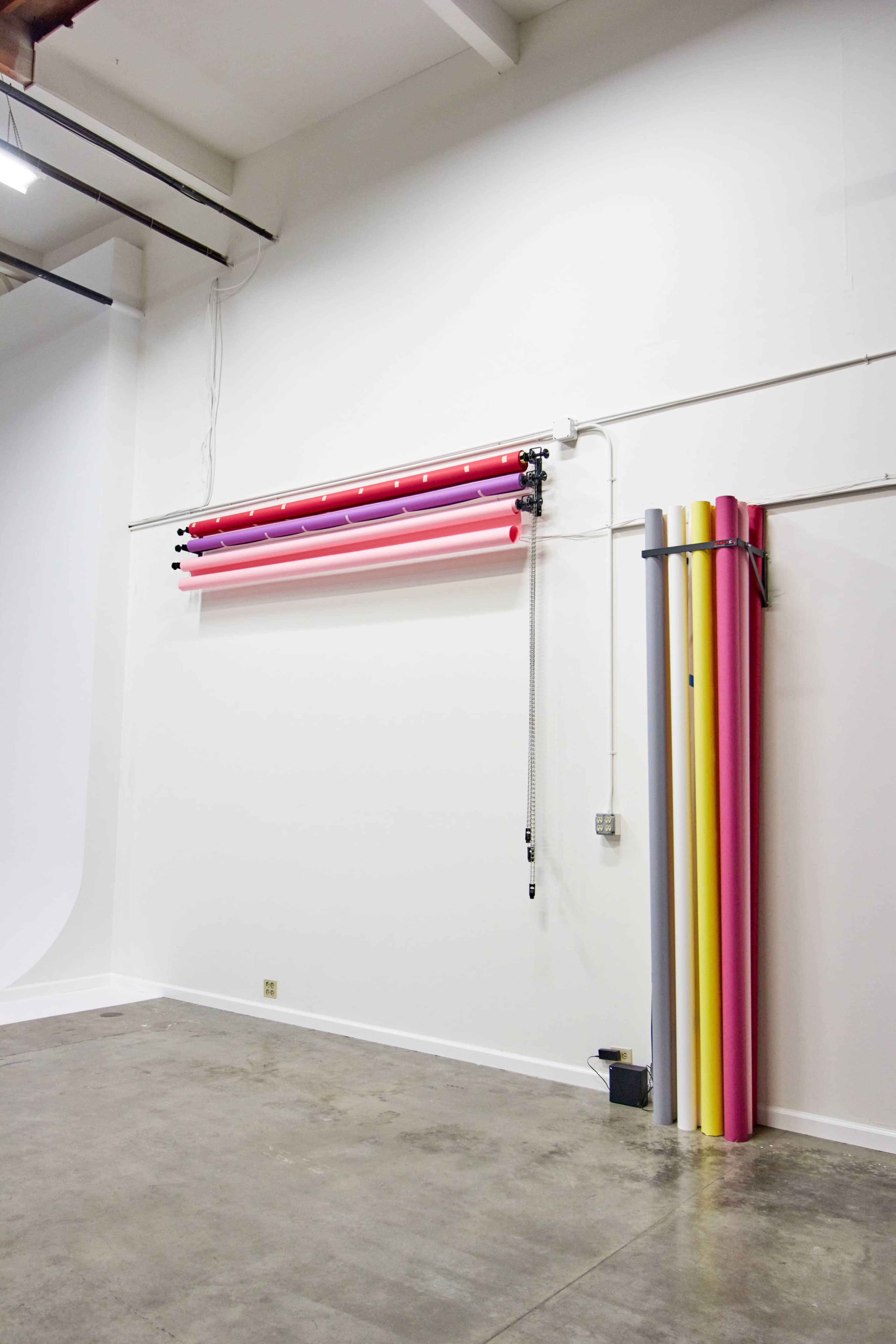 A white wall with rolls of colored backdrop paper hanging above and vertically stacked to the right on the floor.