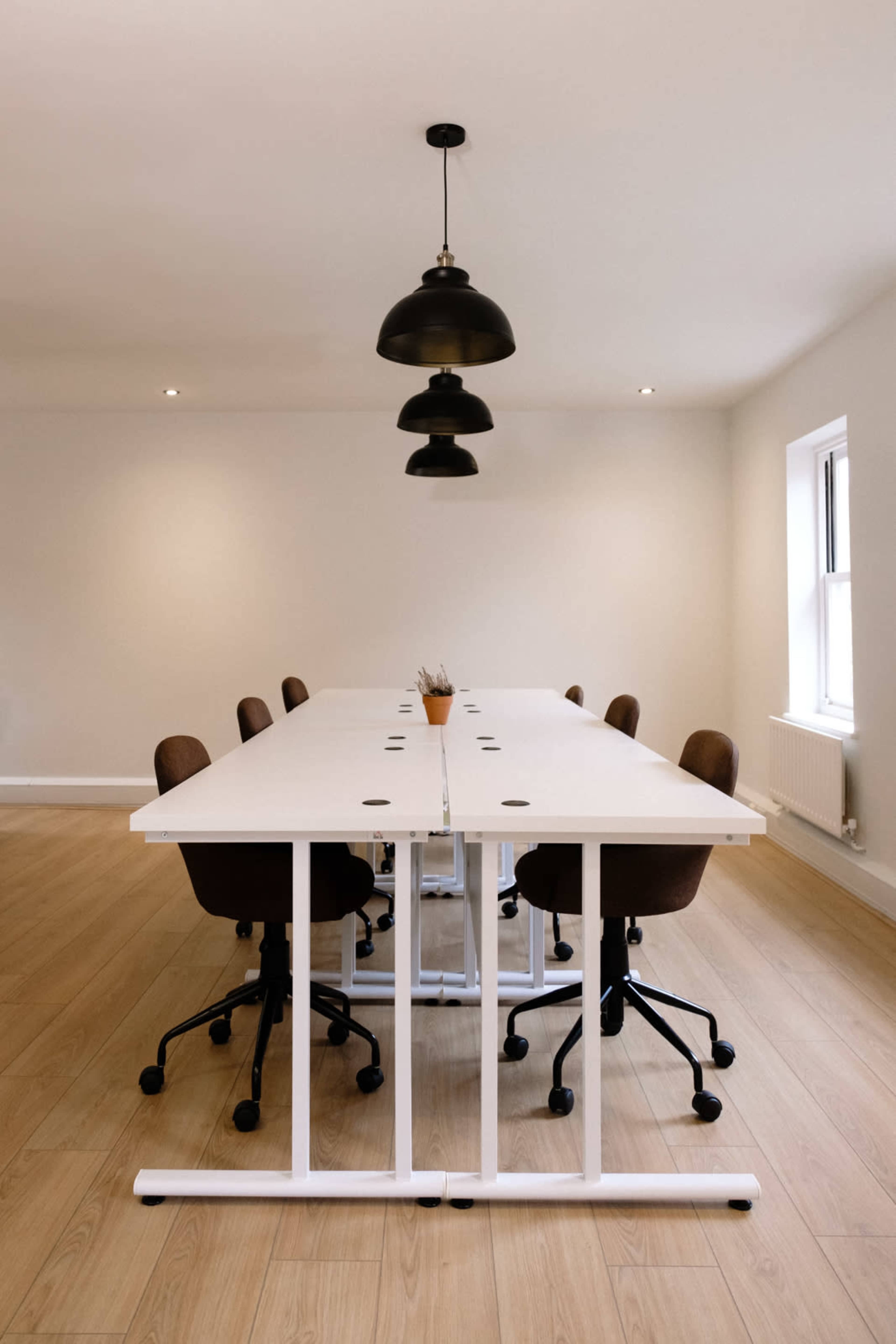 A modern meeting room features a long white table surrounded by brown office chairs, illuminated by black pendant lights.