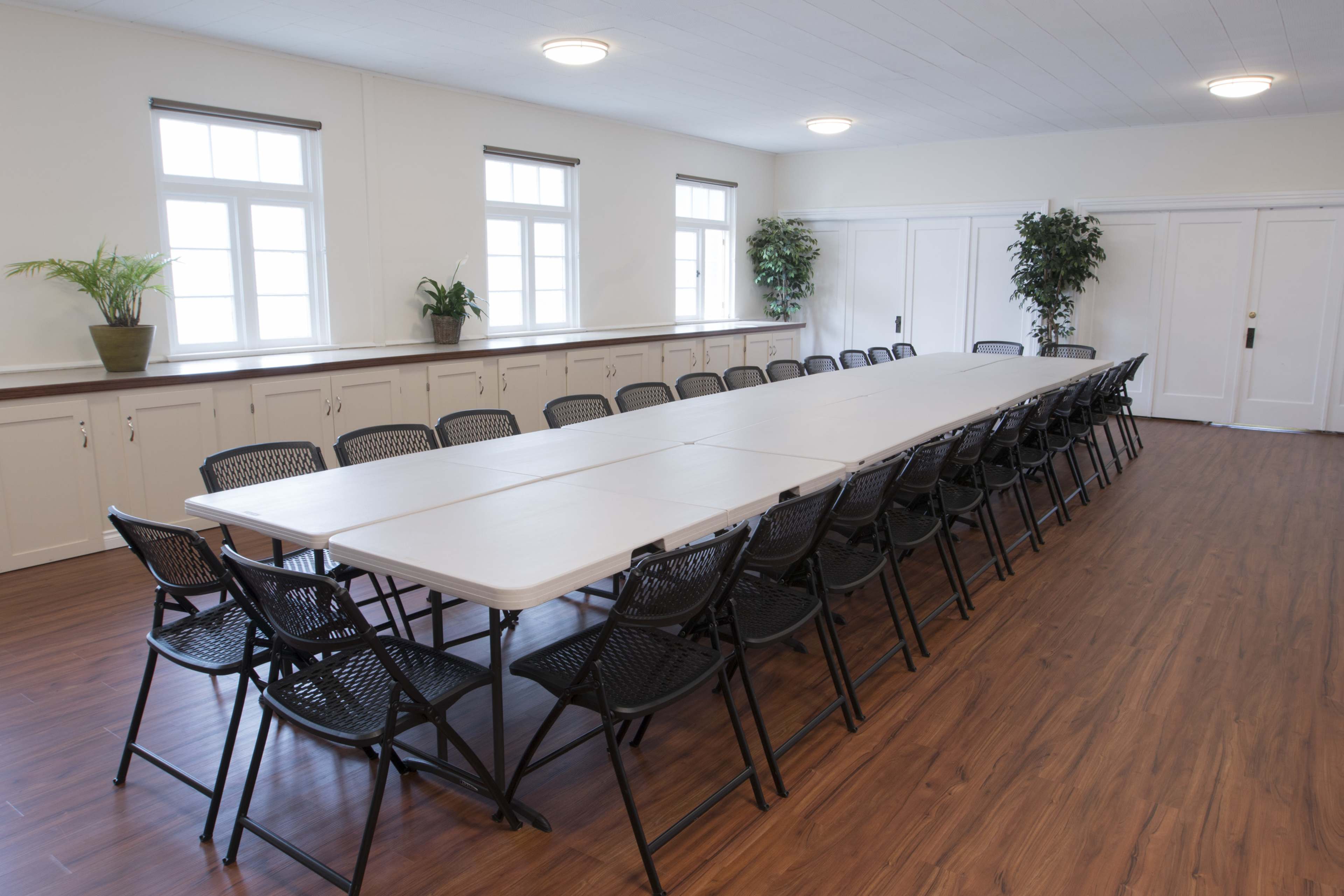 A long, rectangular table surrounded by black folding chairs in a well-lit meeting room with windows and plants.