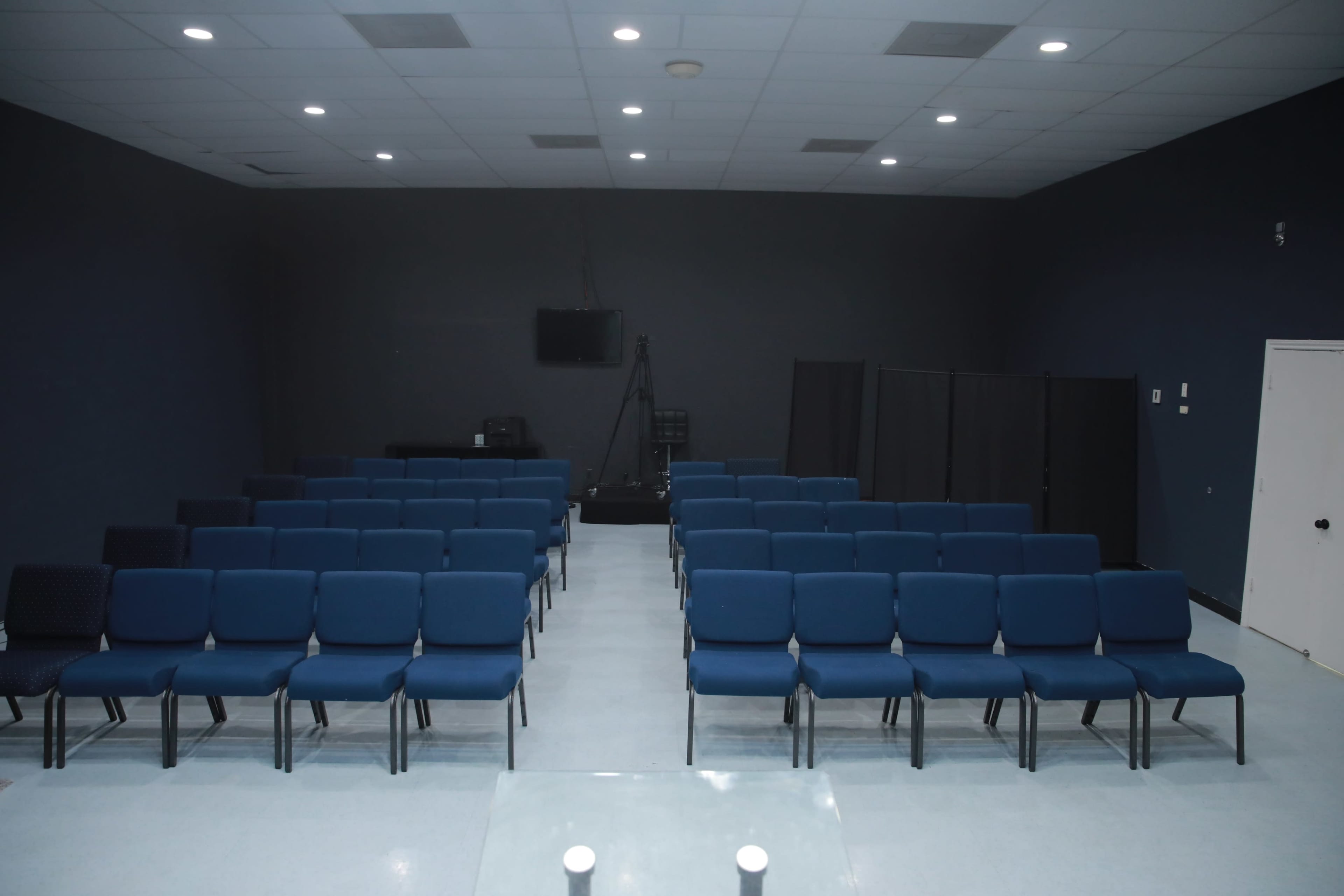 A row of blue chairs is arranged in a conference room with a dark wall and a small setup in the background.
