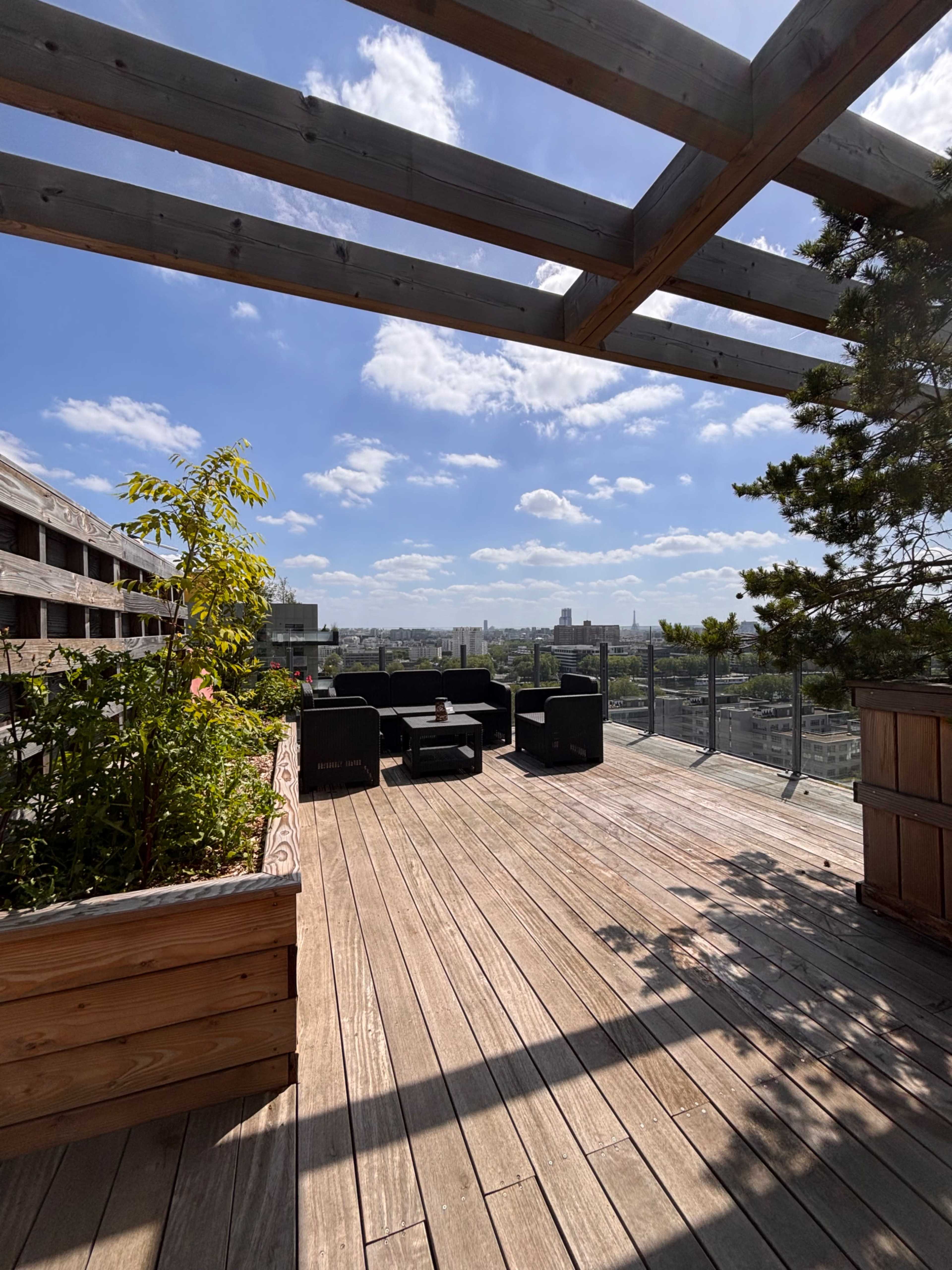 A wooden deck features seating areas and plants, overlooking a city skyline under a partly cloudy sky.