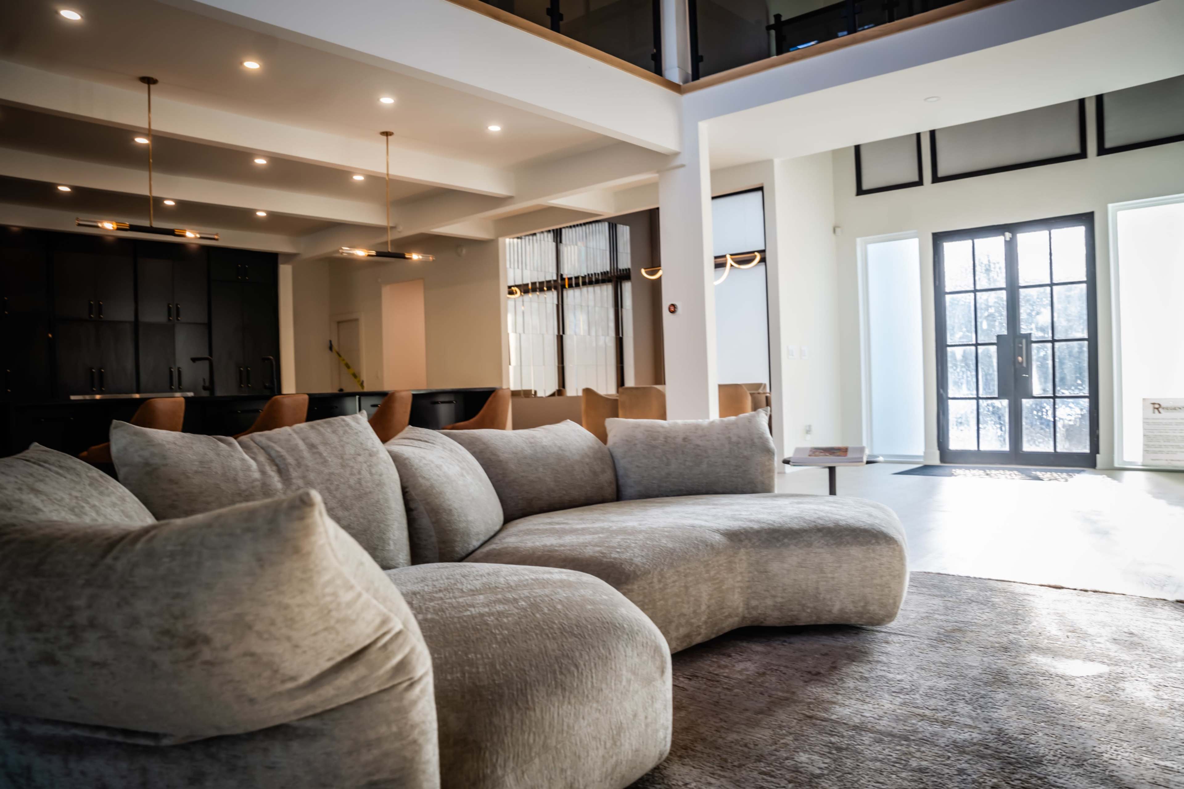 A large, curved gray sofa occupies the foreground of a spacious, modern living area with a black kitchen cabinetry in the background.