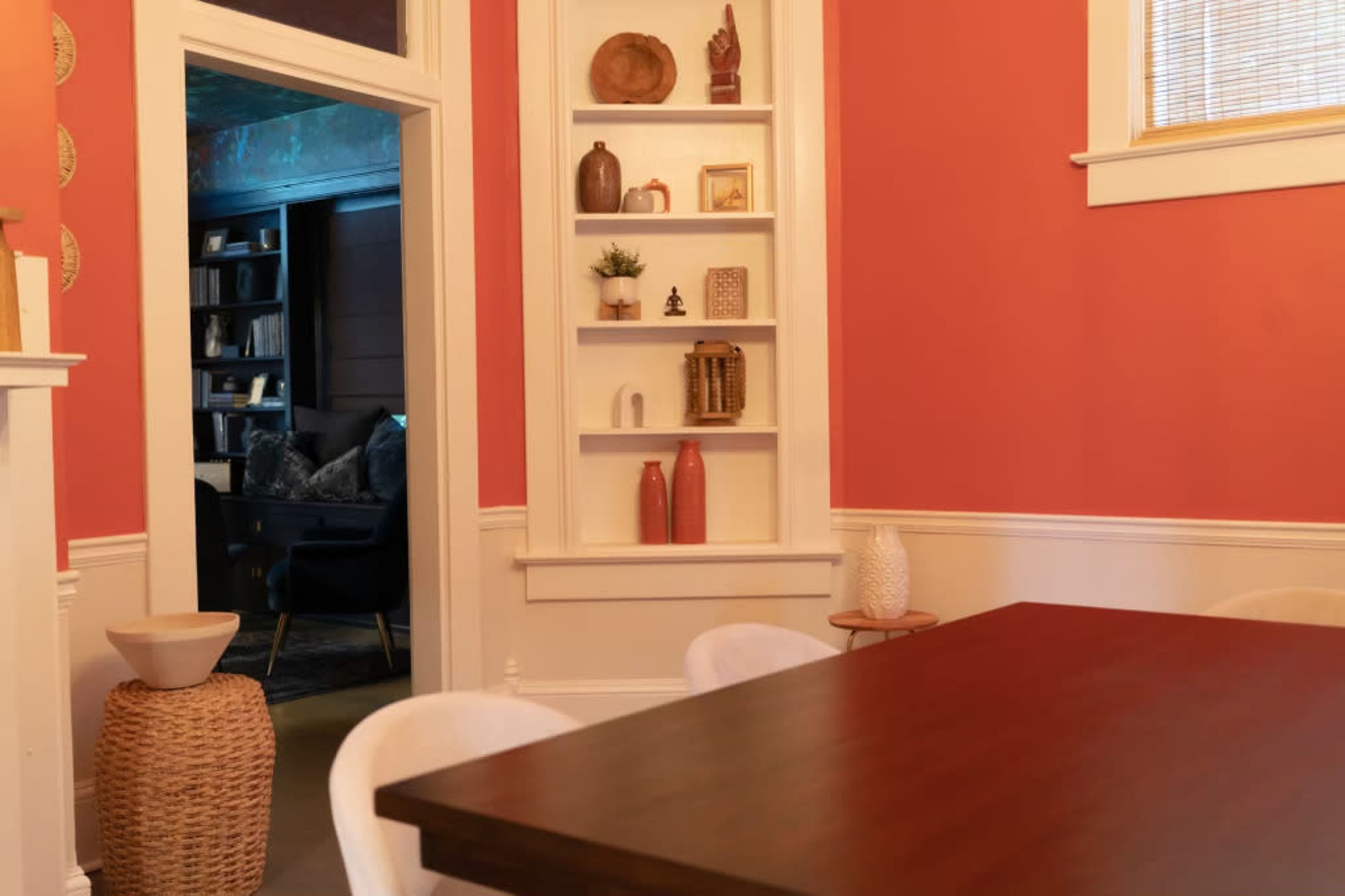 A dining area features a wooden table, surrounded by white chairs, with built-in shelves displaying decorative items against a coral-colored wall.