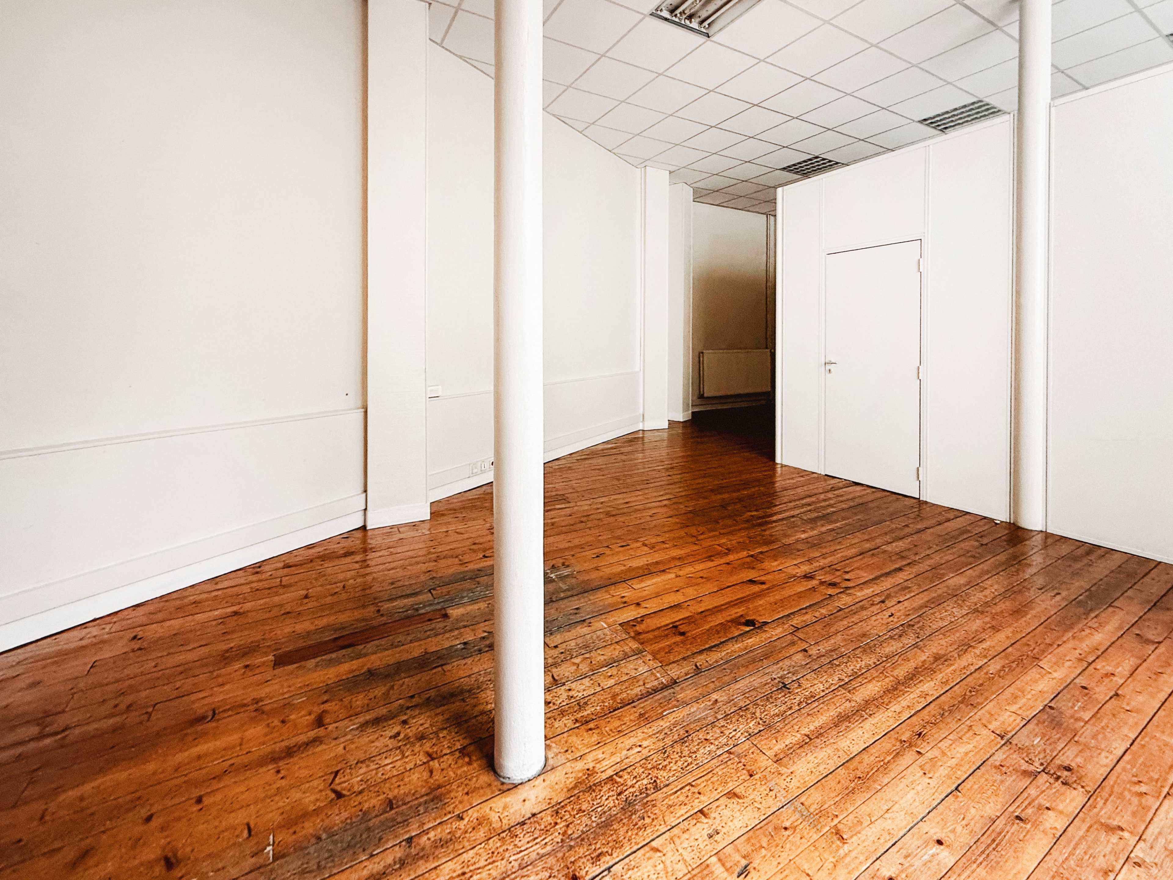 The image shows a spacious, empty room with wooden flooring, a white wall, and a white door on the far side.