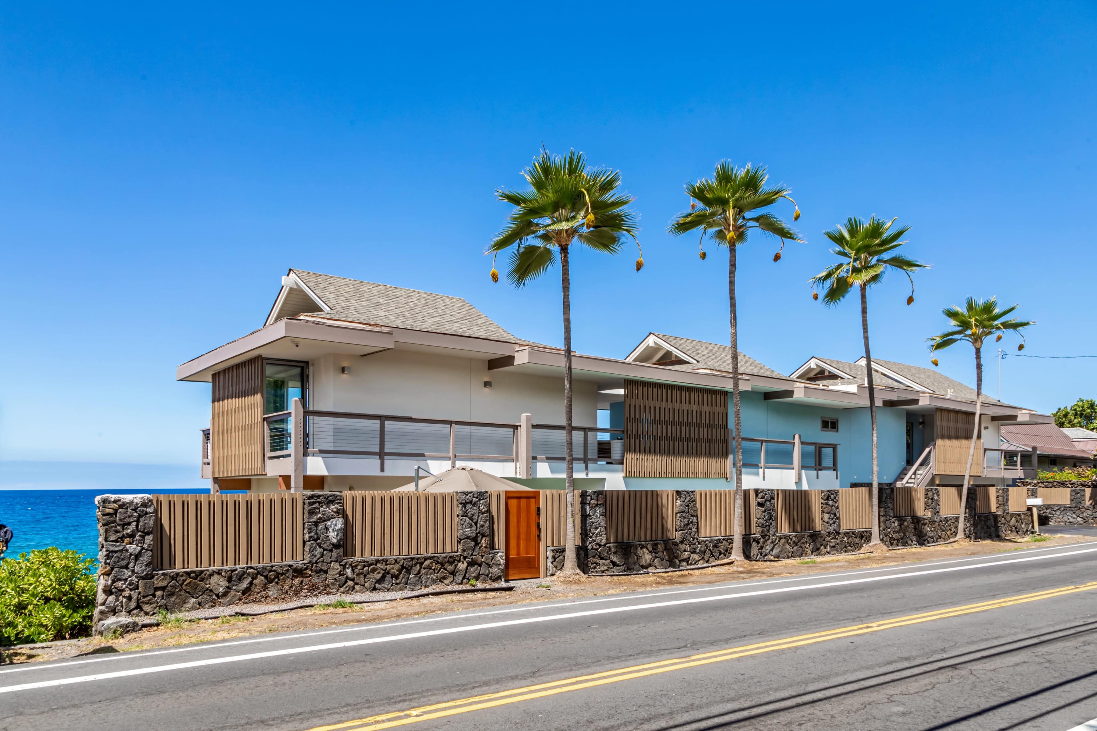 A row of modern homes with palm trees line the roadside next to the ocean.