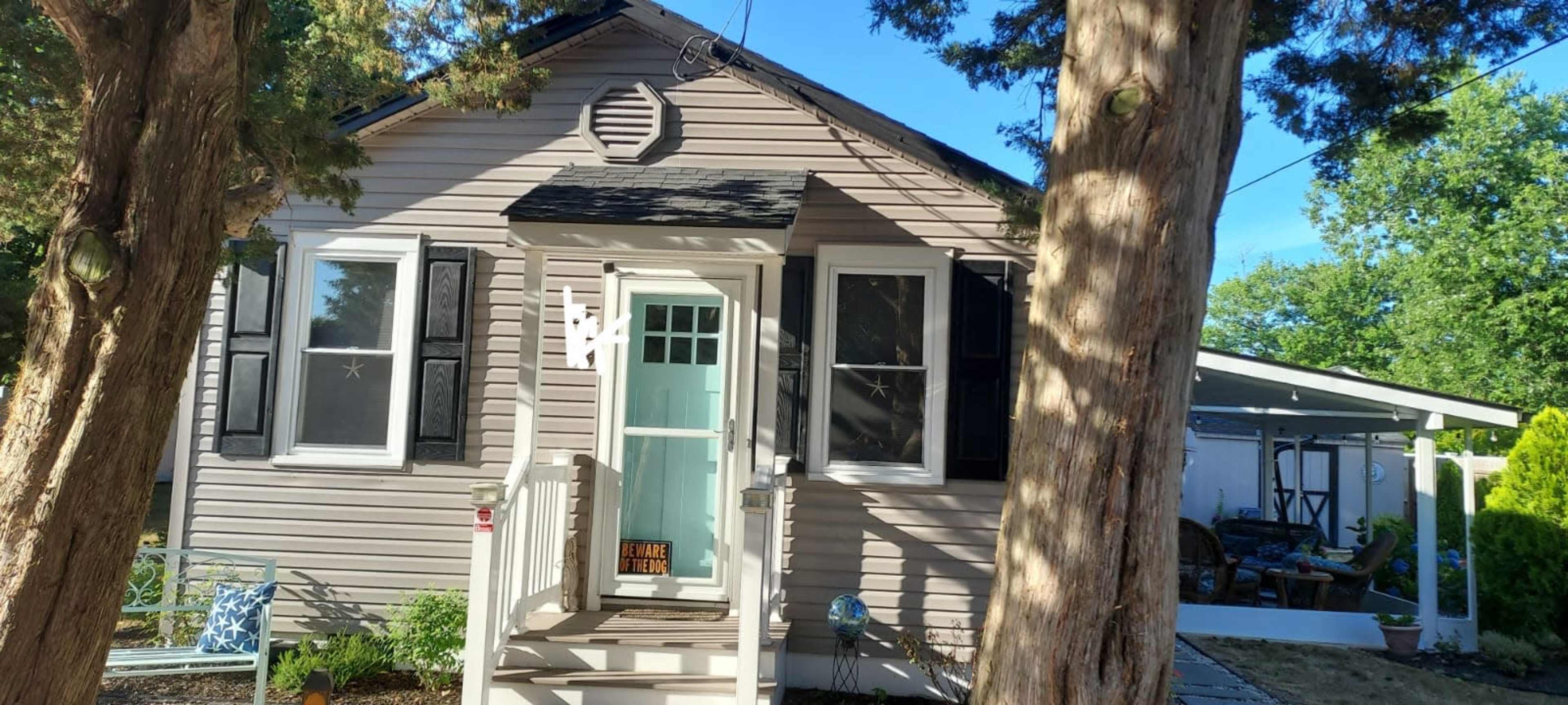A small, single-story house with a light-colored exterior and black shutters is surrounded by trees and features a front porch with a welcoming sign.