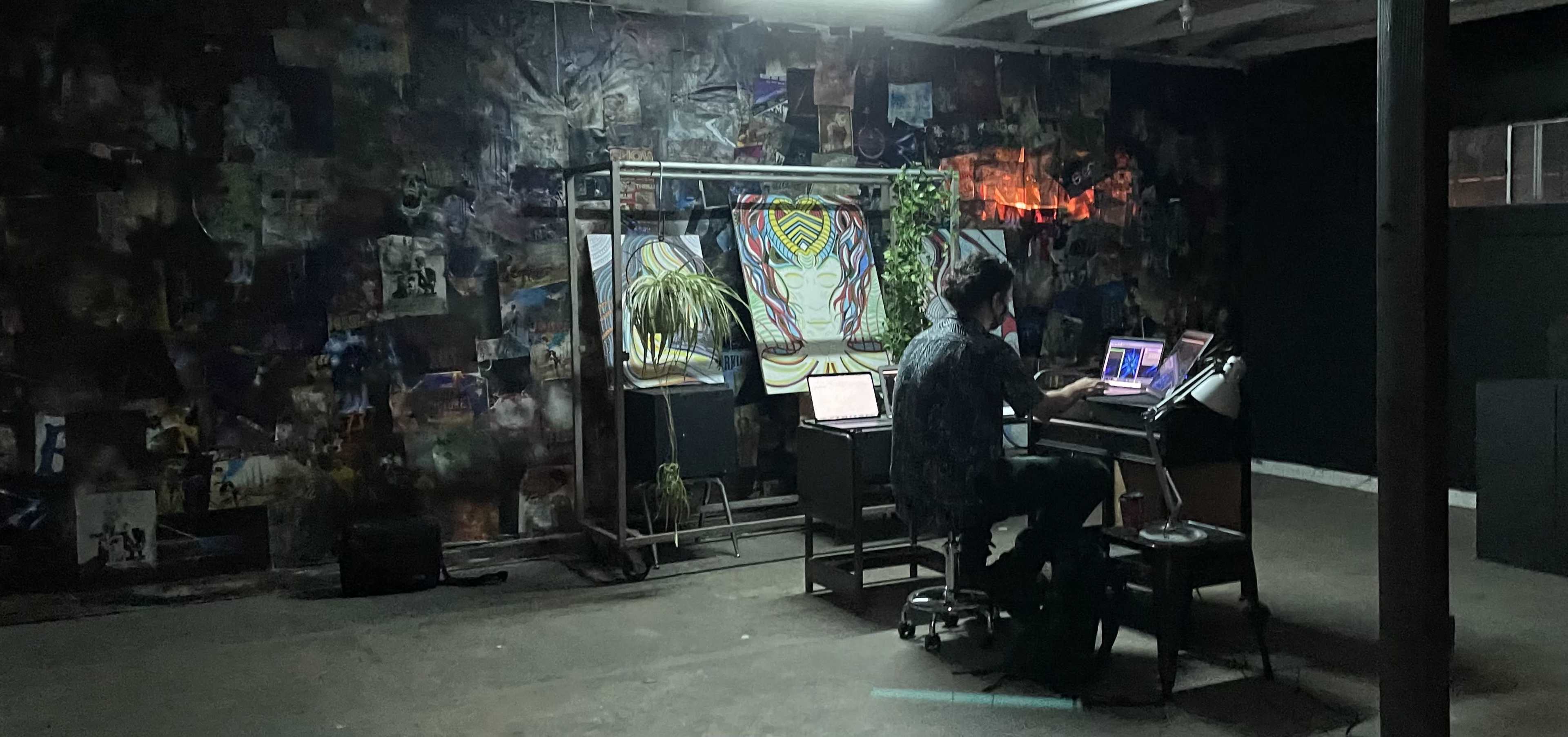 A person sits at a desk in a dimly lit room surrounded by a collage of images on the walls.