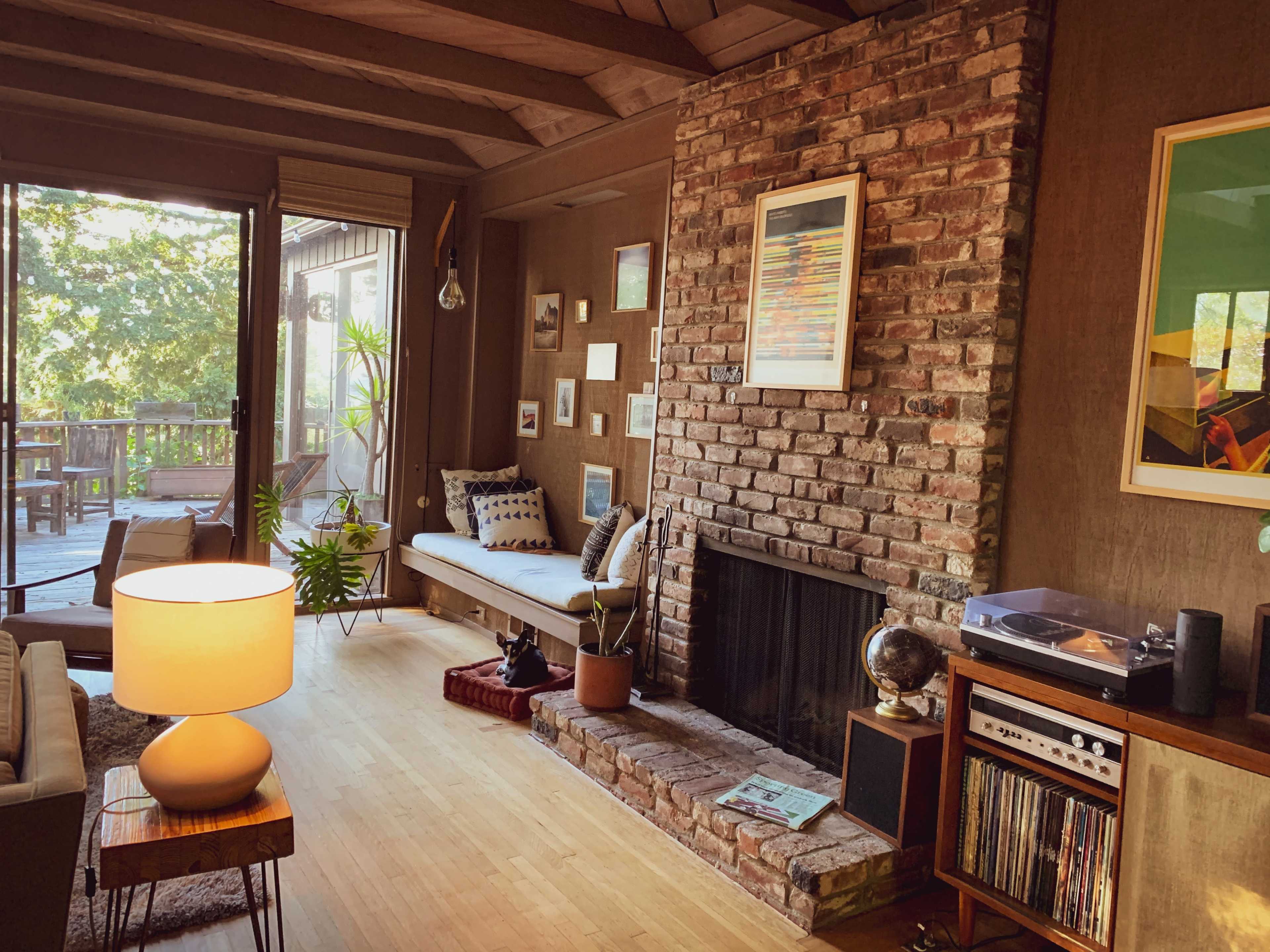 A cozy living room featuring a brick fireplace, wooden beams, and a record player beside a large window with a view of a deck.