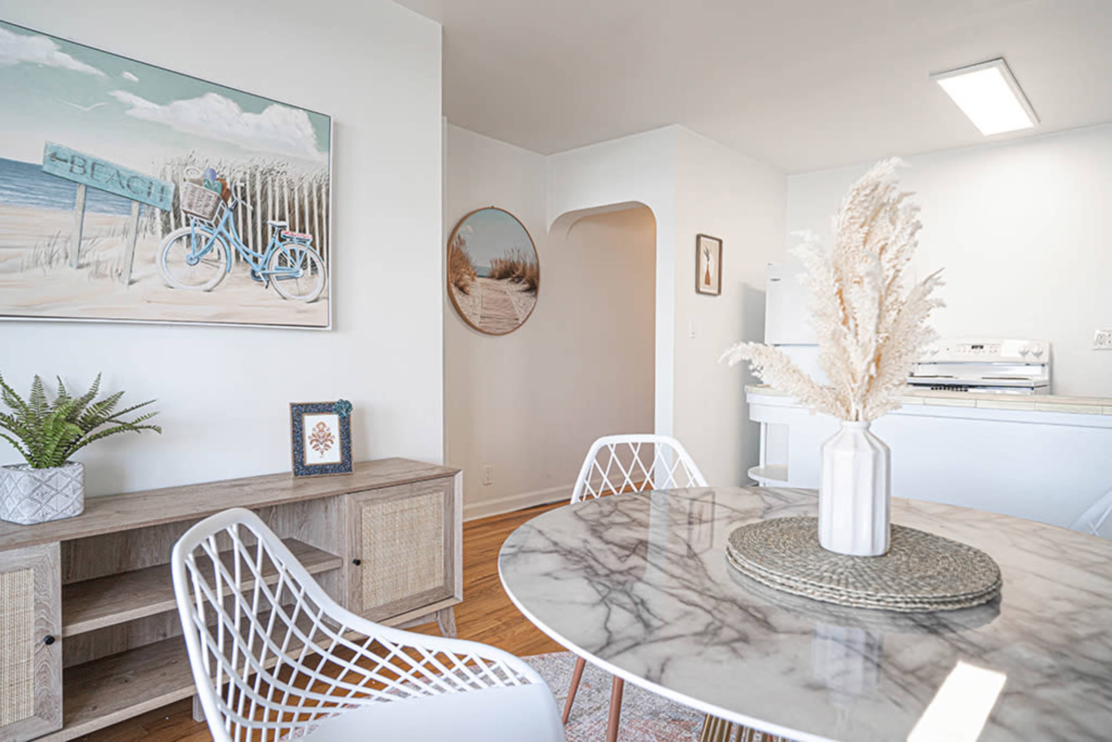 A dining area with a round marble table surrounded by white chairs, featuring a coastal-themed painting and a decorative plant.
