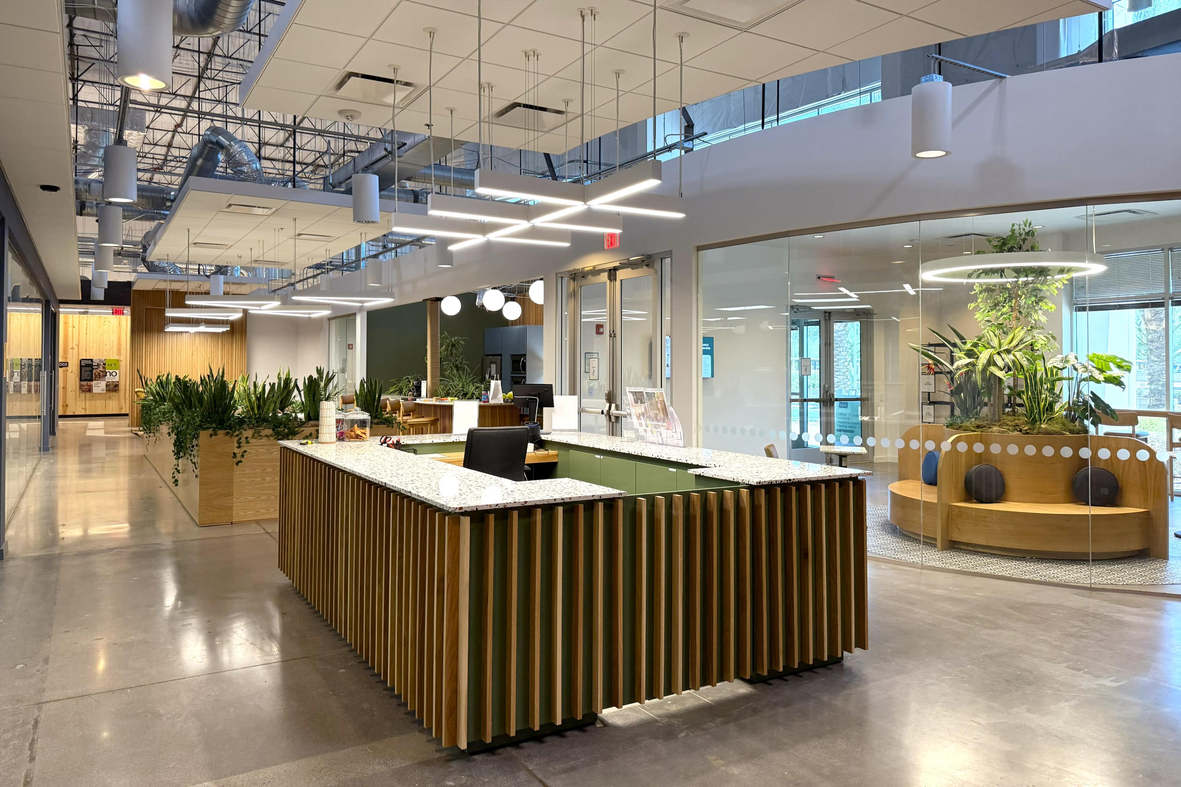 The image shows a modern office reception area with a wooden front desk and seating spaces surrounded by large plants.