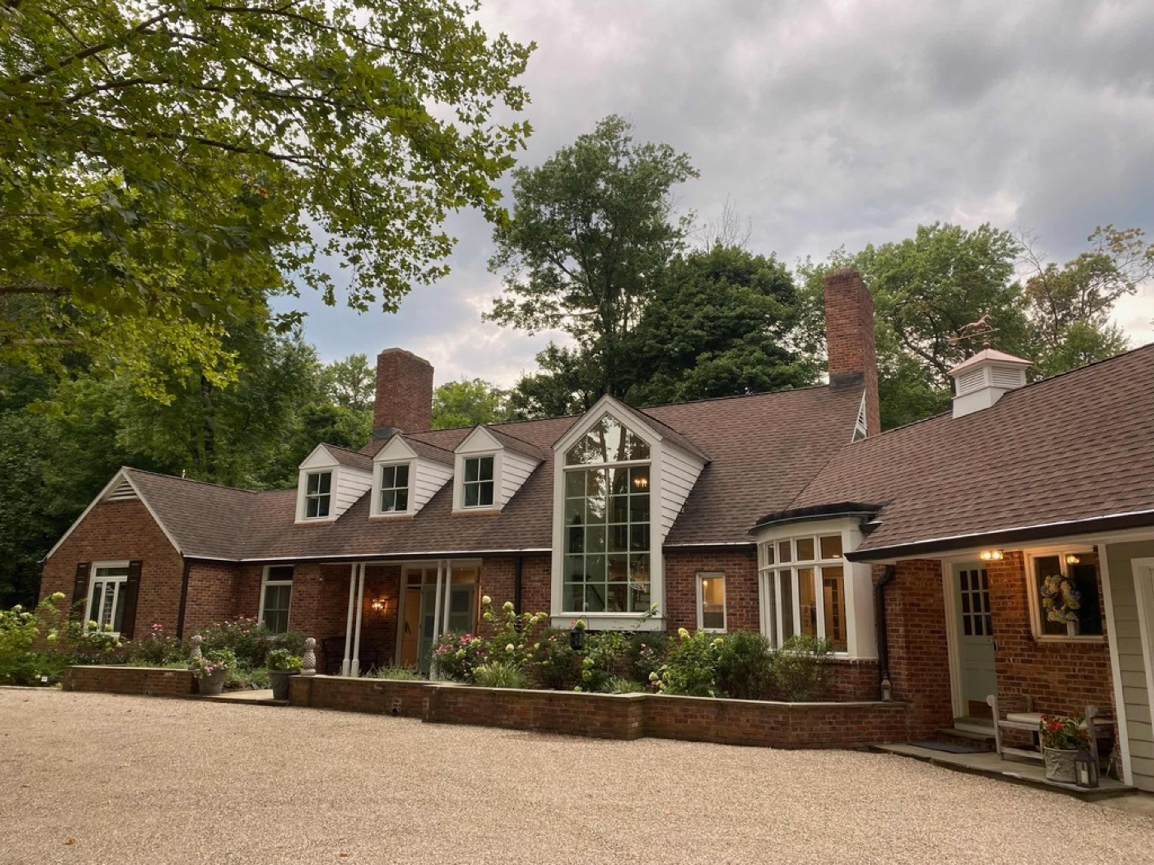 A large brick house with multiple gables and a central window is surrounded by trees and a gravel driveway.