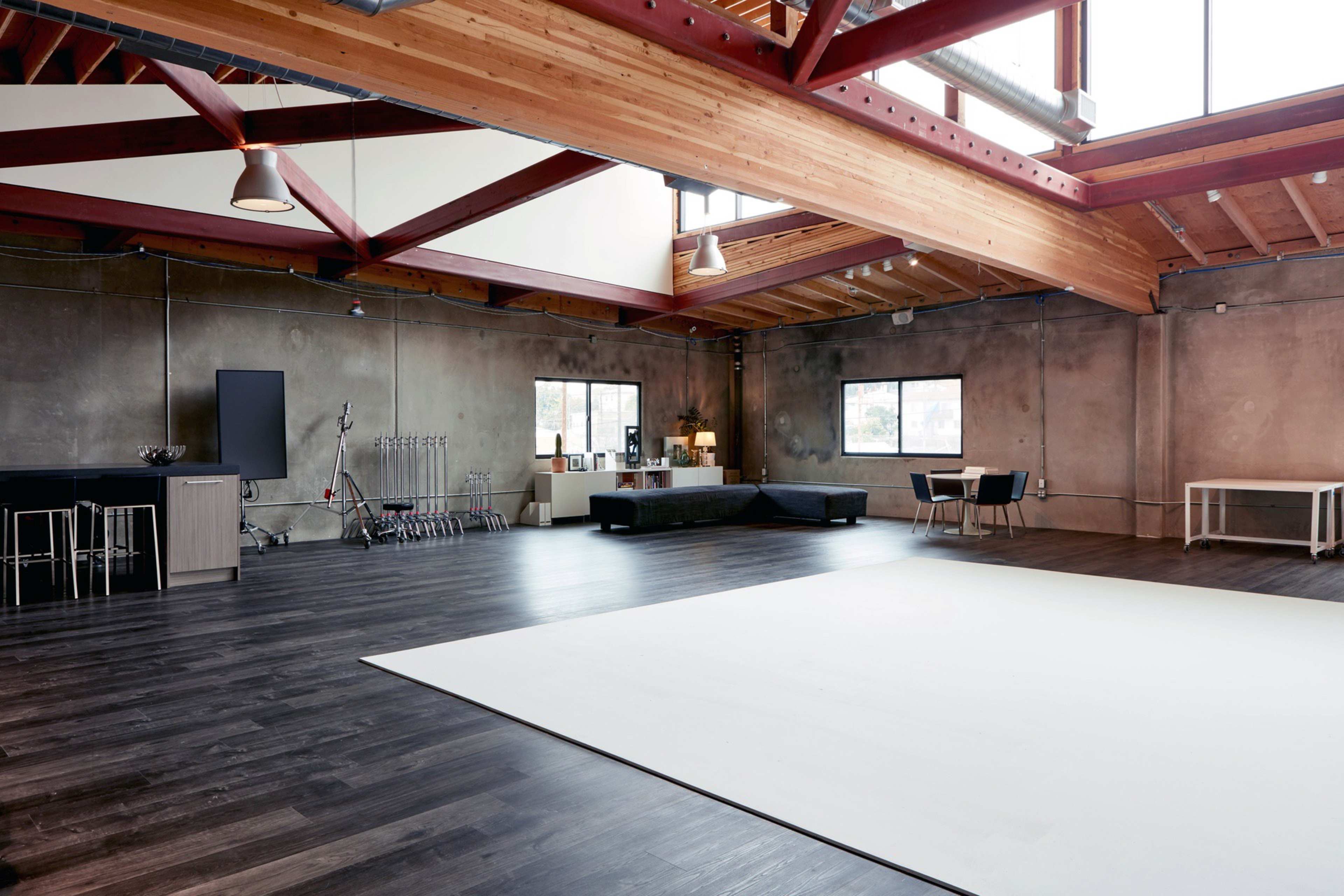 A spacious, industrial-style room with high ceilings, wooden beams, and large windows, featuring a mix of furniture and an empty white area on the floor.