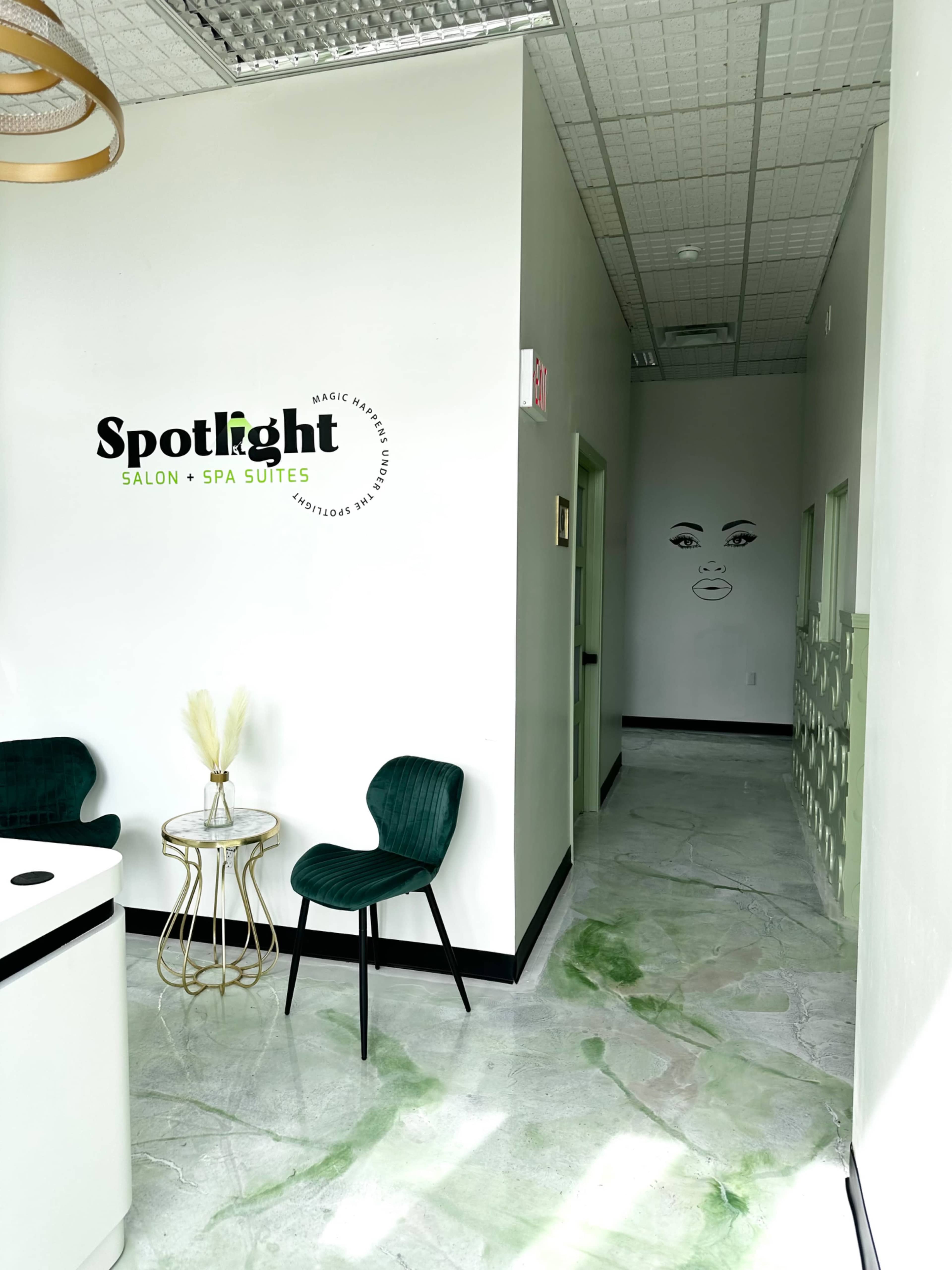 The image shows a modern salon and spa entrance featuring a green accent chair, a small round table, and a corridor leading to more private suites with a stylized facial mural on the wall.