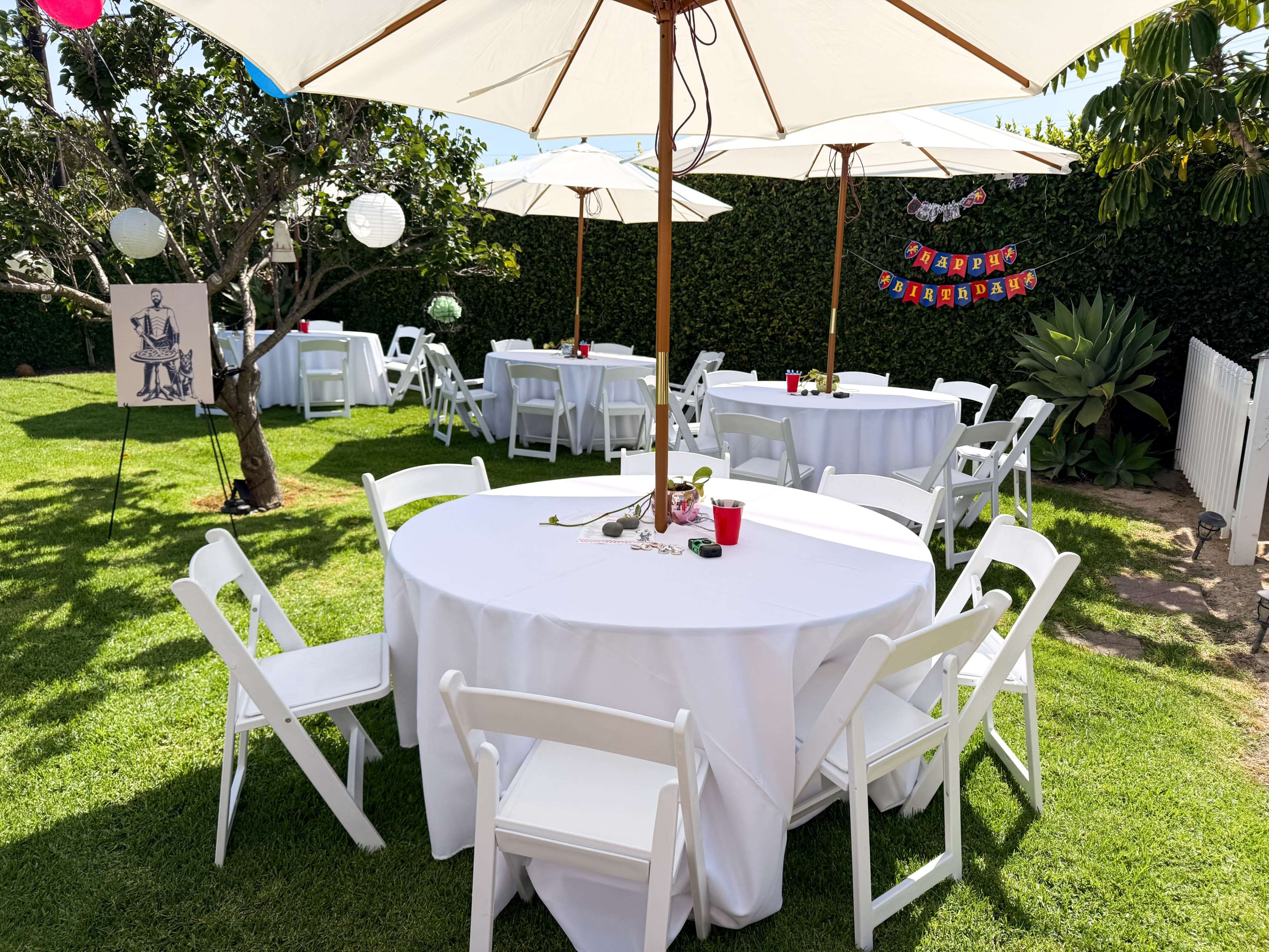 The image shows a decorated outdoor space with white tables and chairs under large umbrellas, accompanied by a tree, festive decorations, and a banner.
