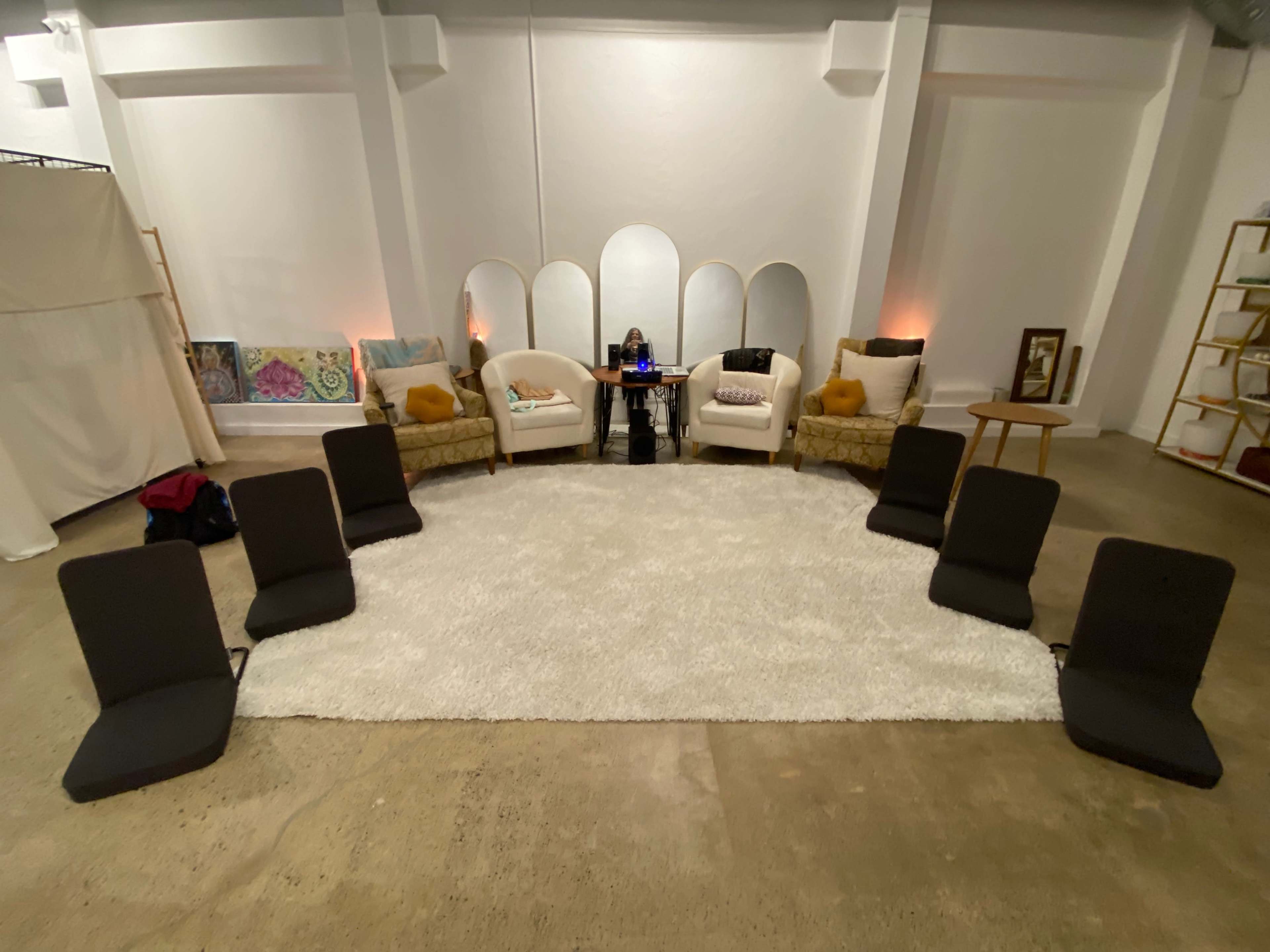 The image shows a spacious room arranged with two armchairs and a table in the center, surrounded by several floor chairs on a soft white rug.