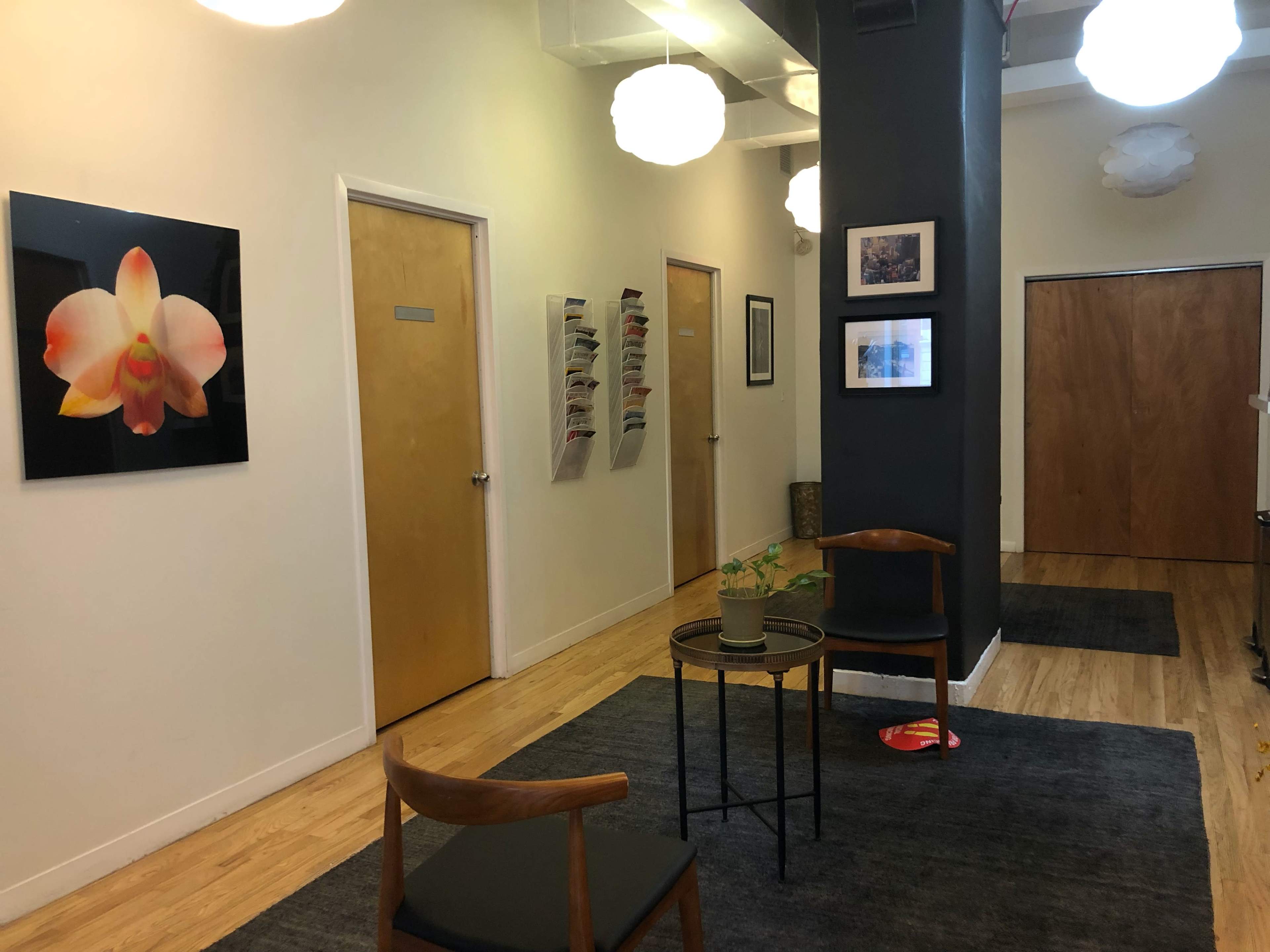 A hallway with wooden flooring, featuring doors on either side, a plant on a small table, and a wall adorned with artwork and magazines.
