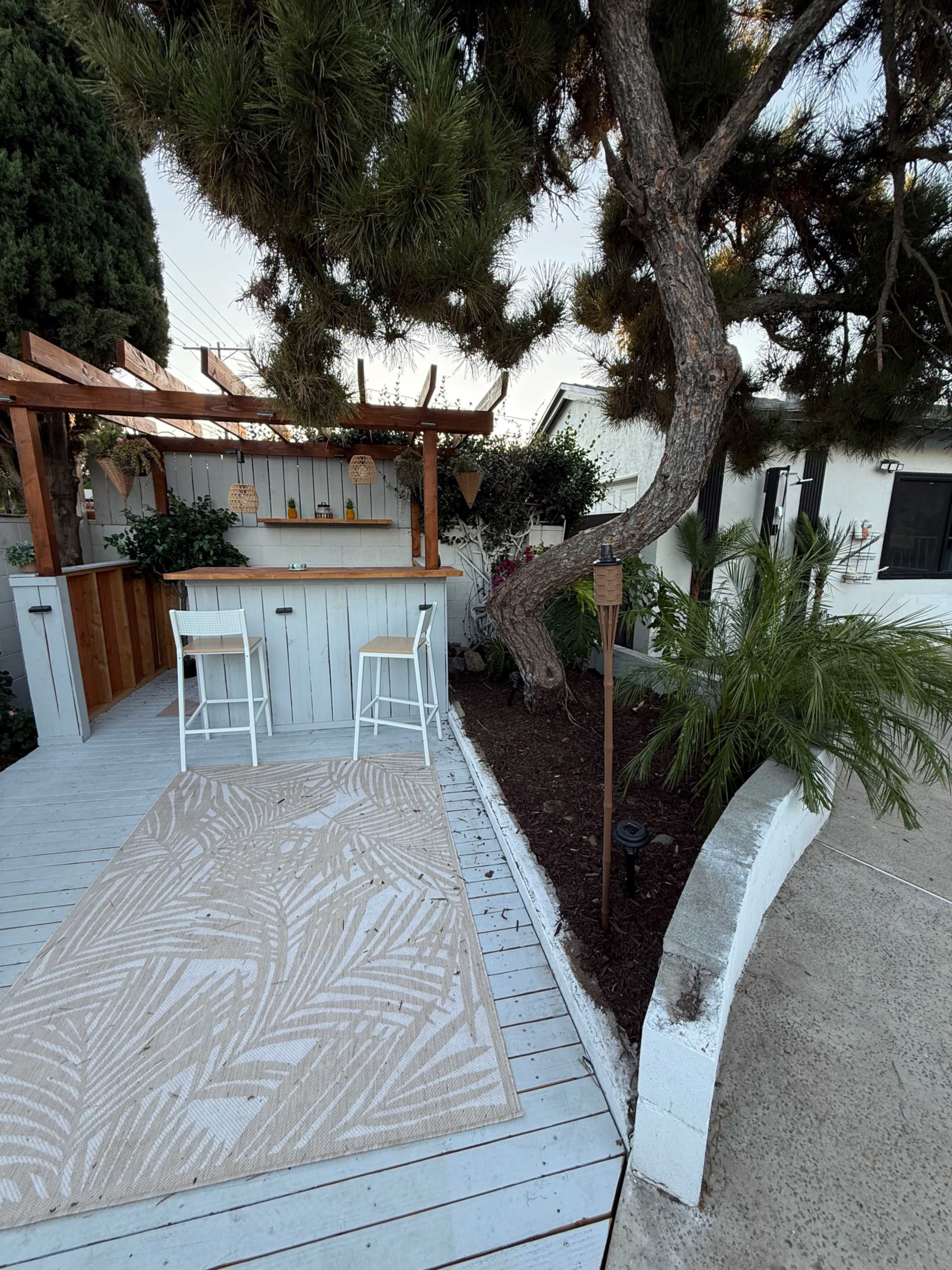 The image shows a small outdoor space featuring a wooden patio with two chairs and a bar area, surrounded by greenery and a tree.