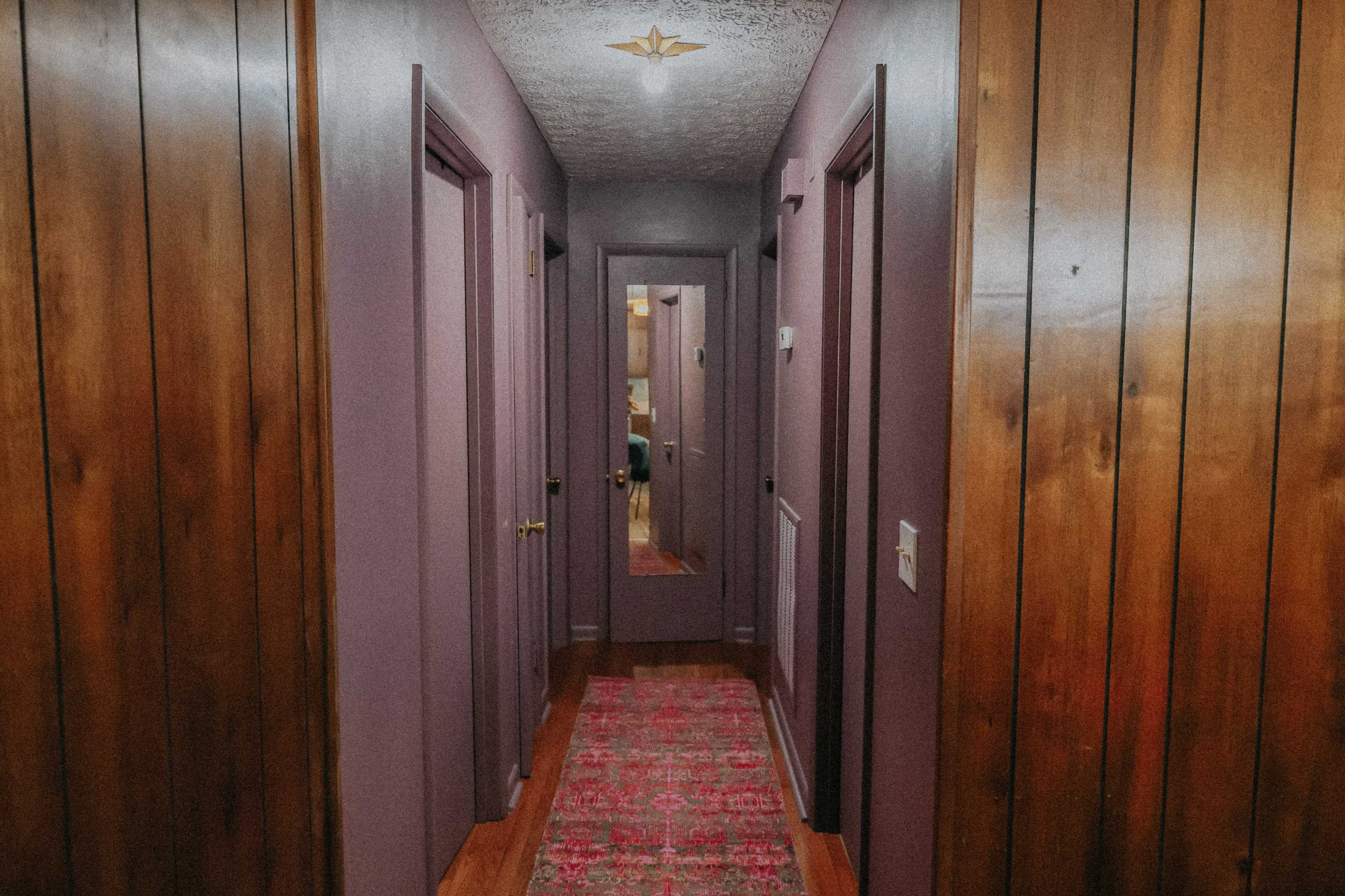 The image shows a hallway with purple walls, wooden paneling, and a patterned rug leading to a mirrored door at the end.