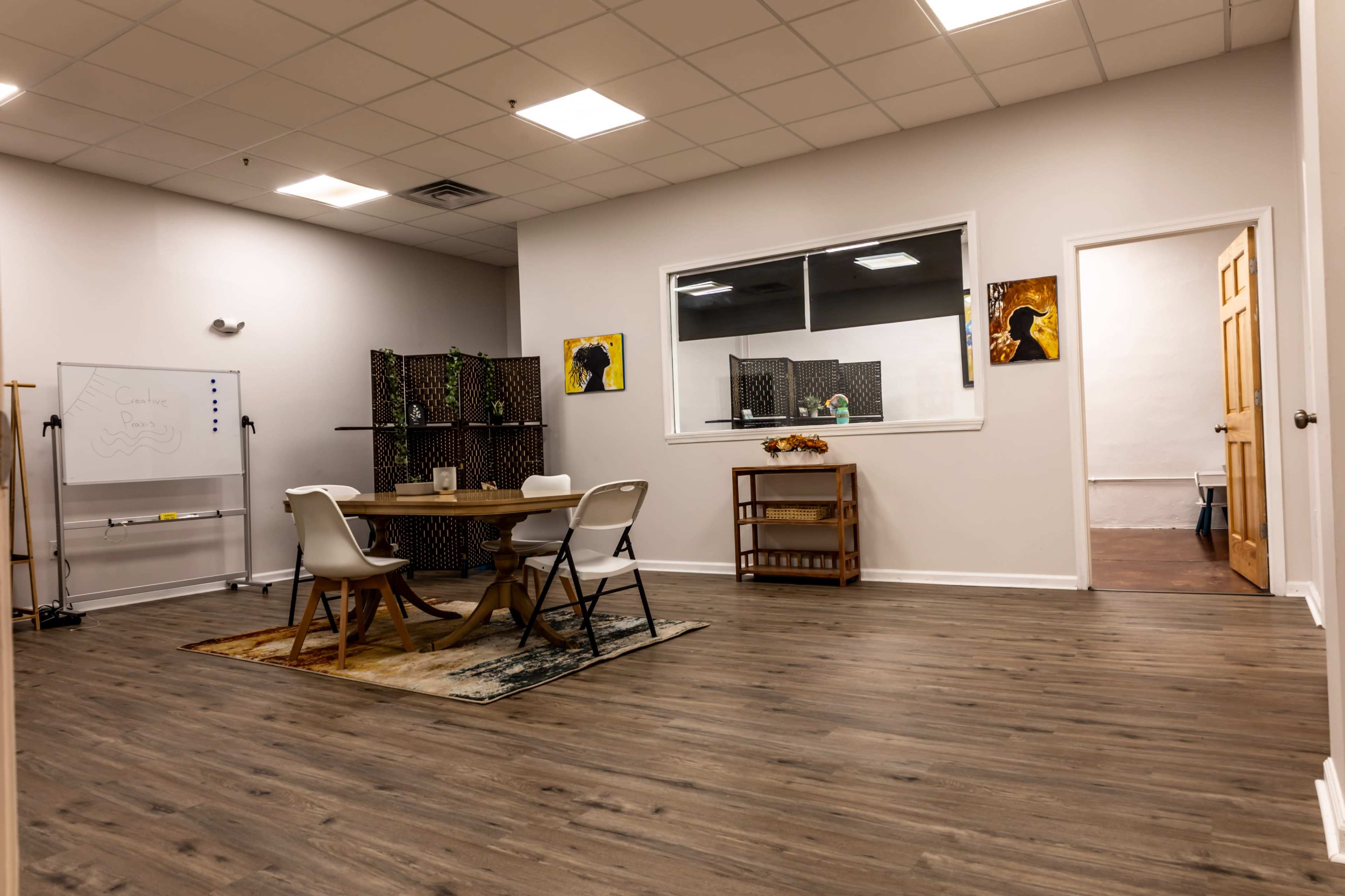 The image shows a spacious room with wooden flooring, featuring a round table surrounded by chairs, a whiteboard, and two artworks hung on the walls.