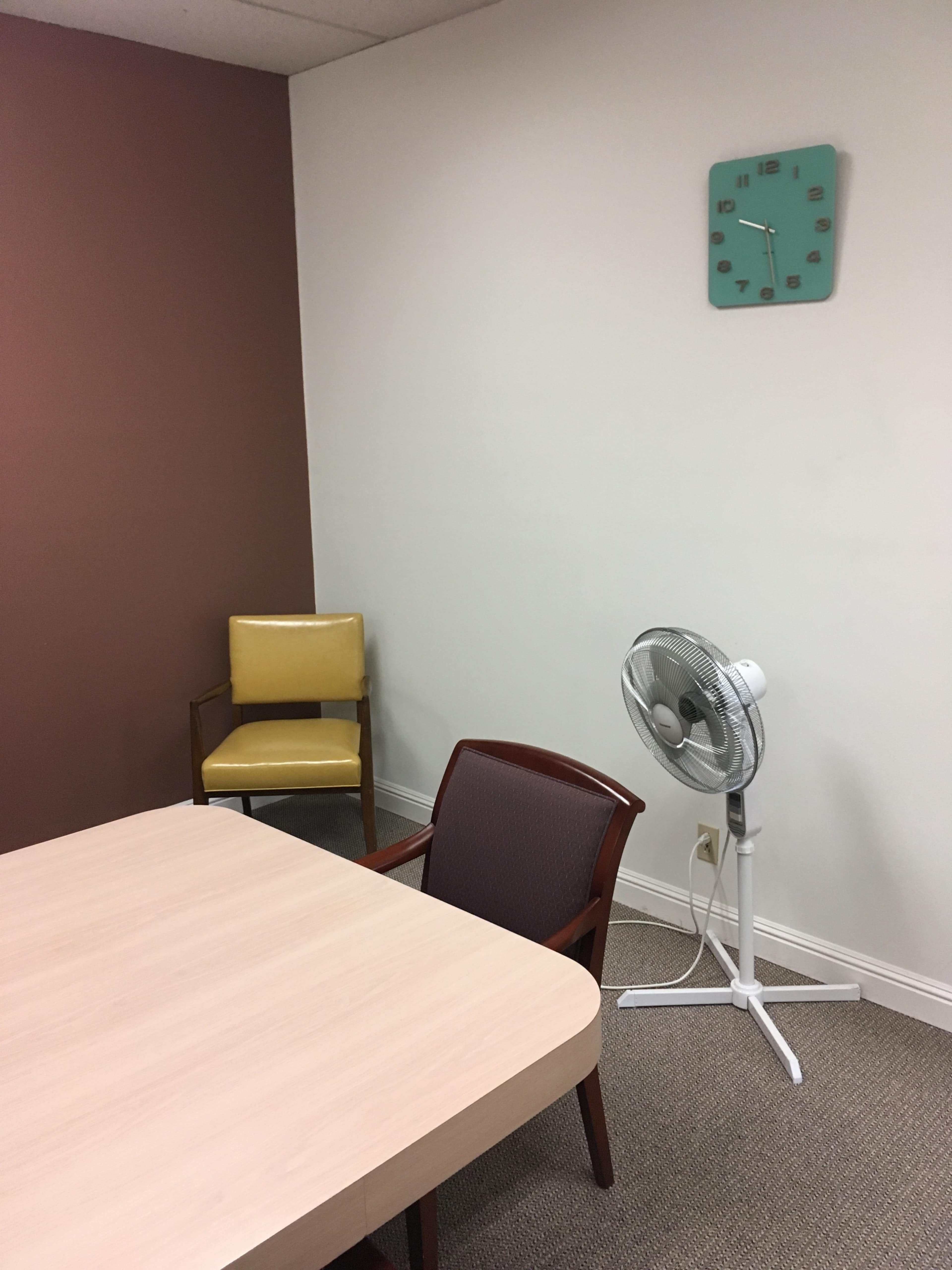 A small office space with a wooden table, two chairs, a wall clock, and a standing fan.