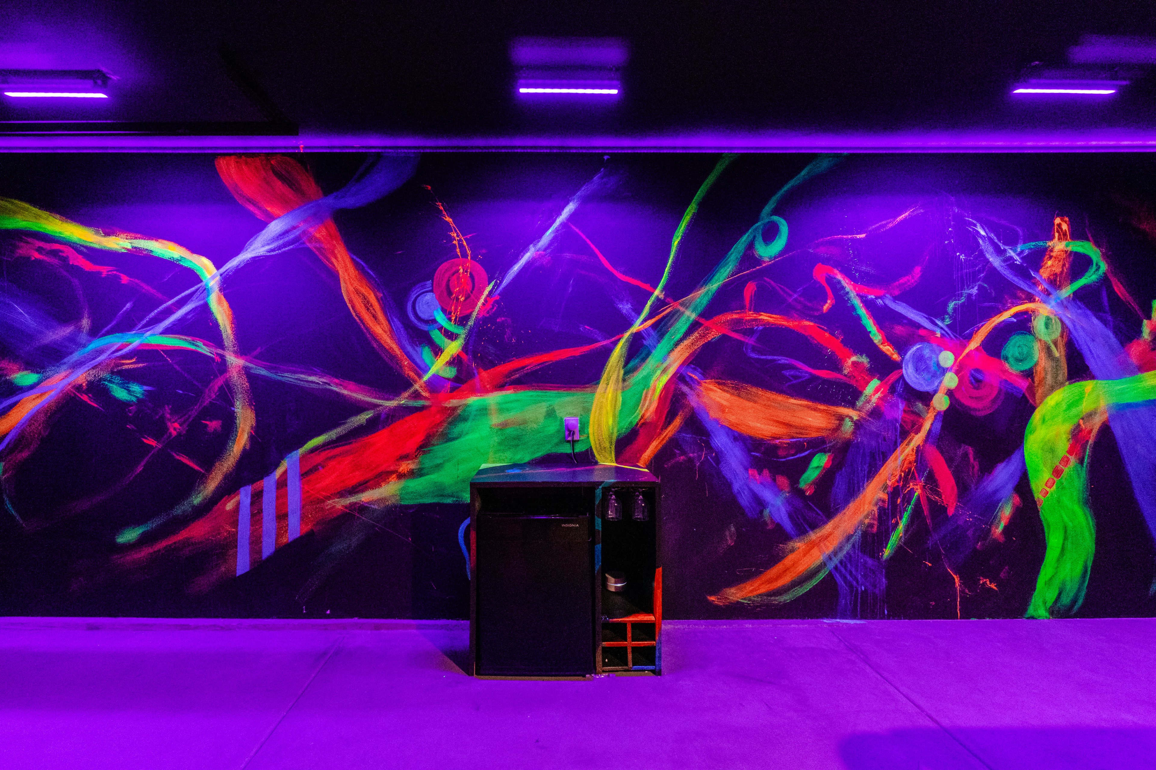 A black wall covered with vibrant, abstract patterns of neon colors and a dark cabinet positioned in front.