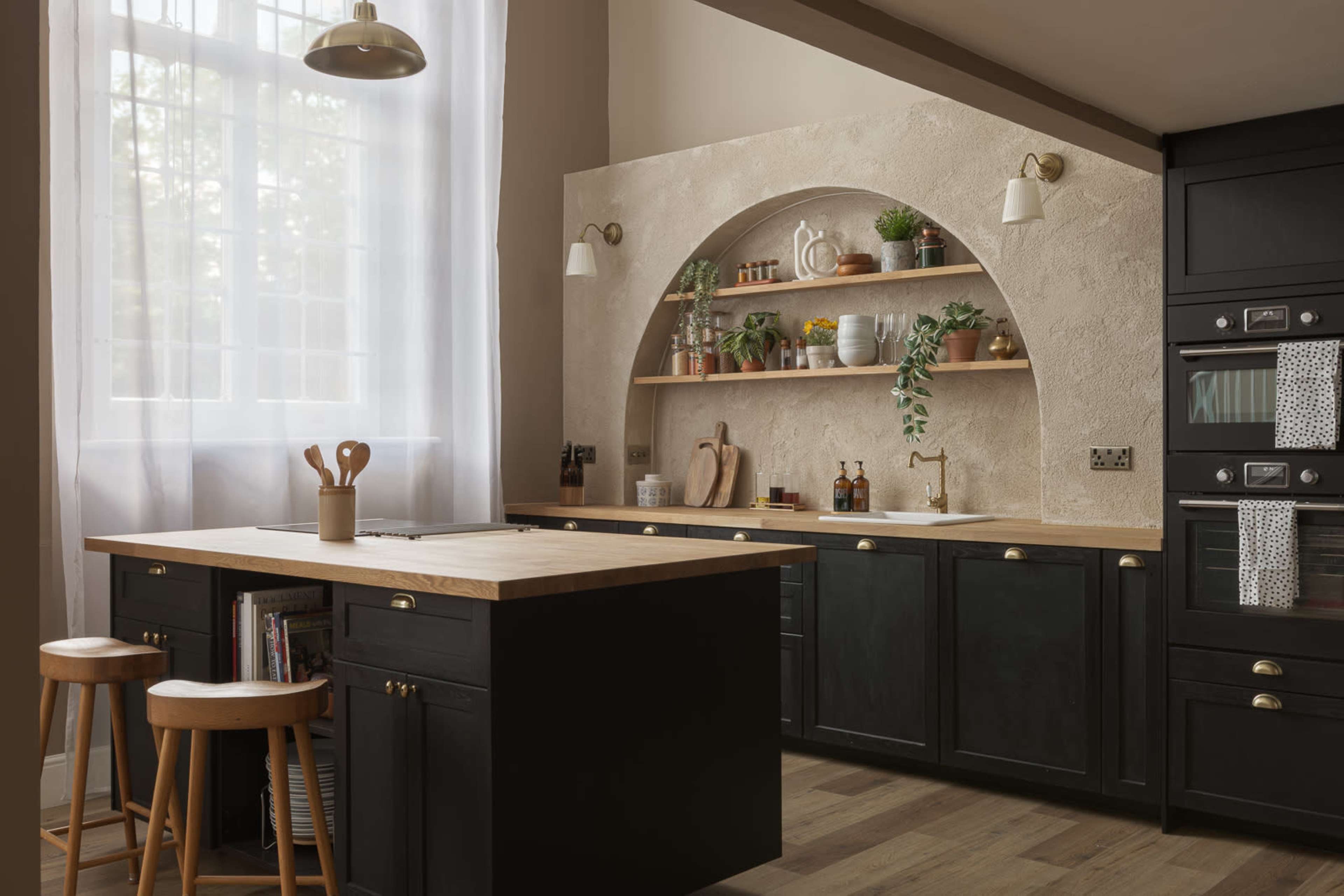 The image shows a modern kitchen featuring a central island with wooden countertops, black cabinetry, and an arched wall shelf adorned with plants and kitchenware.
