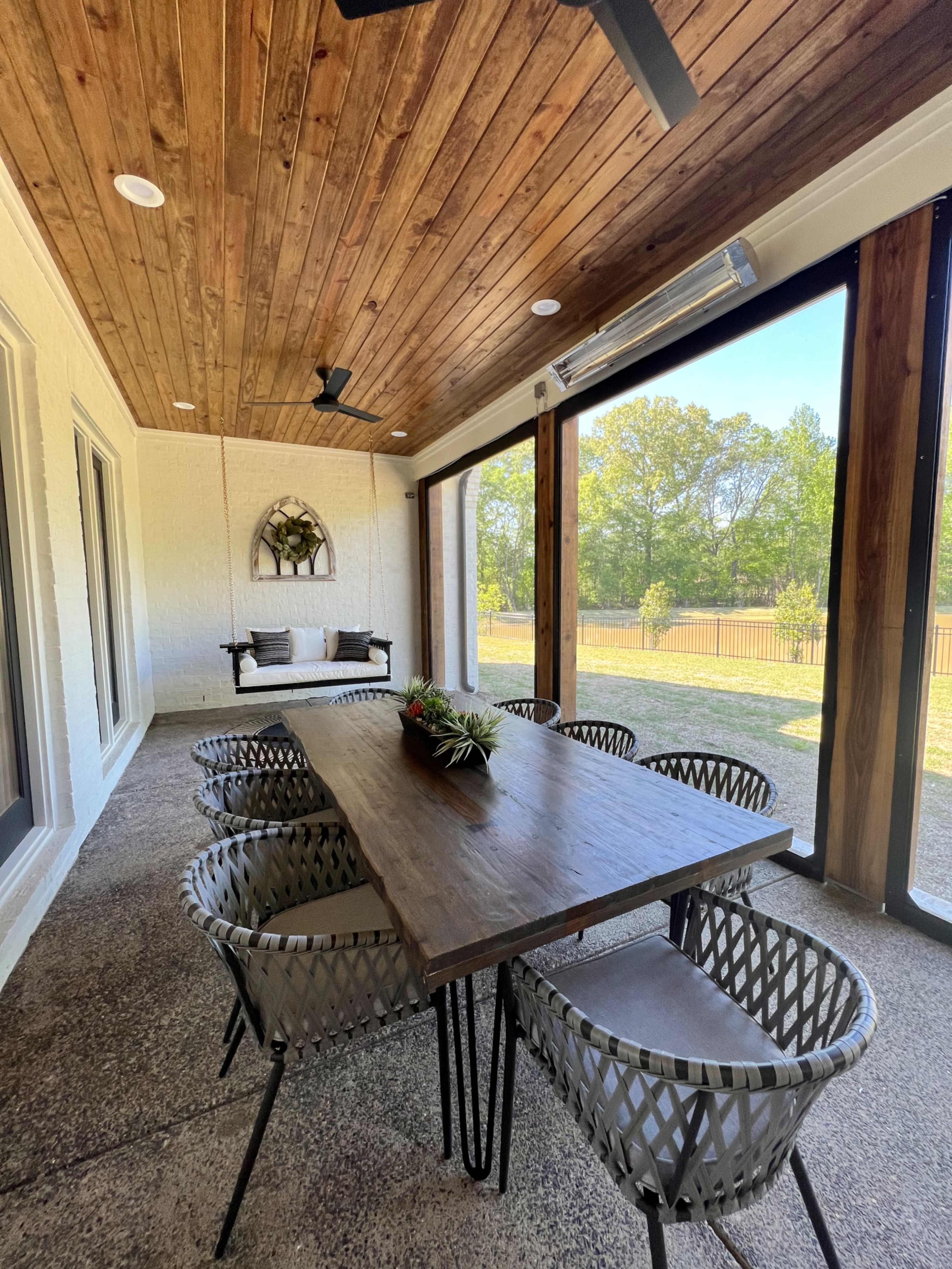 A covered patio features a wooden table surrounded by six woven chairs, with large glass panels and greenery visible outside.