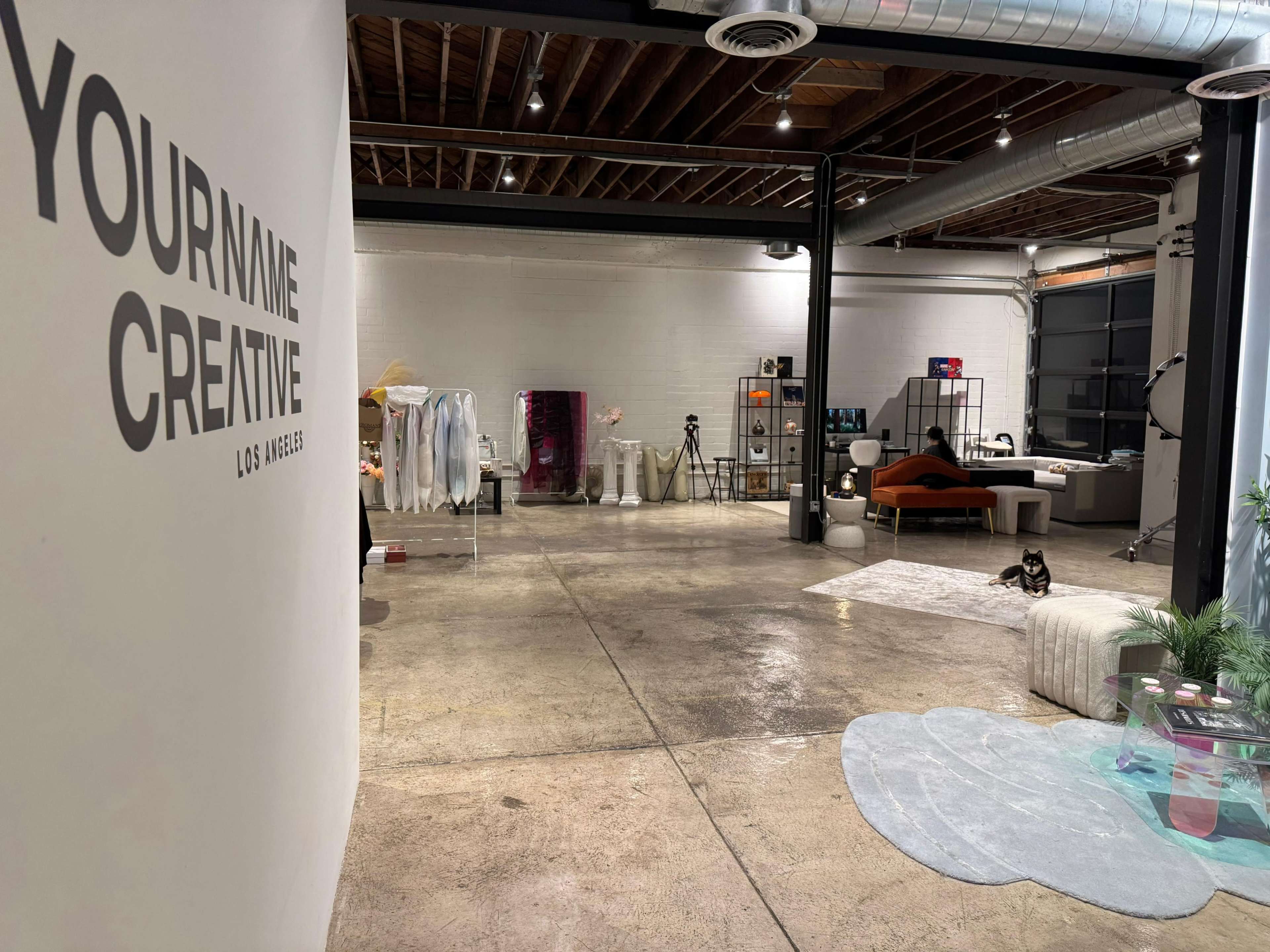 The image shows a spacious studio with a decorative wall featuring the text "YOURNAME CREATIVE," and various items including clothing racks, furniture, and a cat in the foreground.