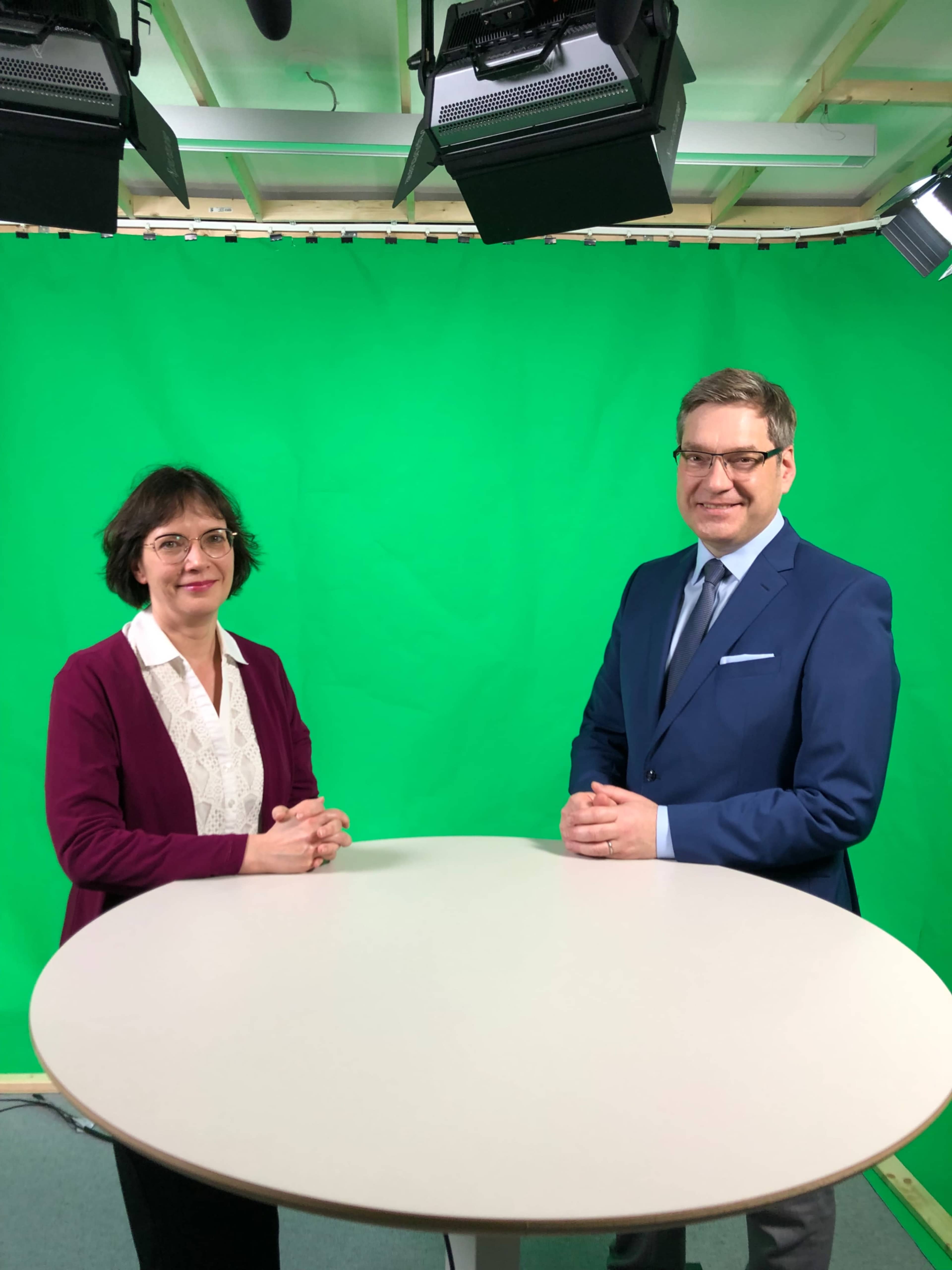 A man and a woman are standing beside a round table in front of a green screen, both looking towards the camera.