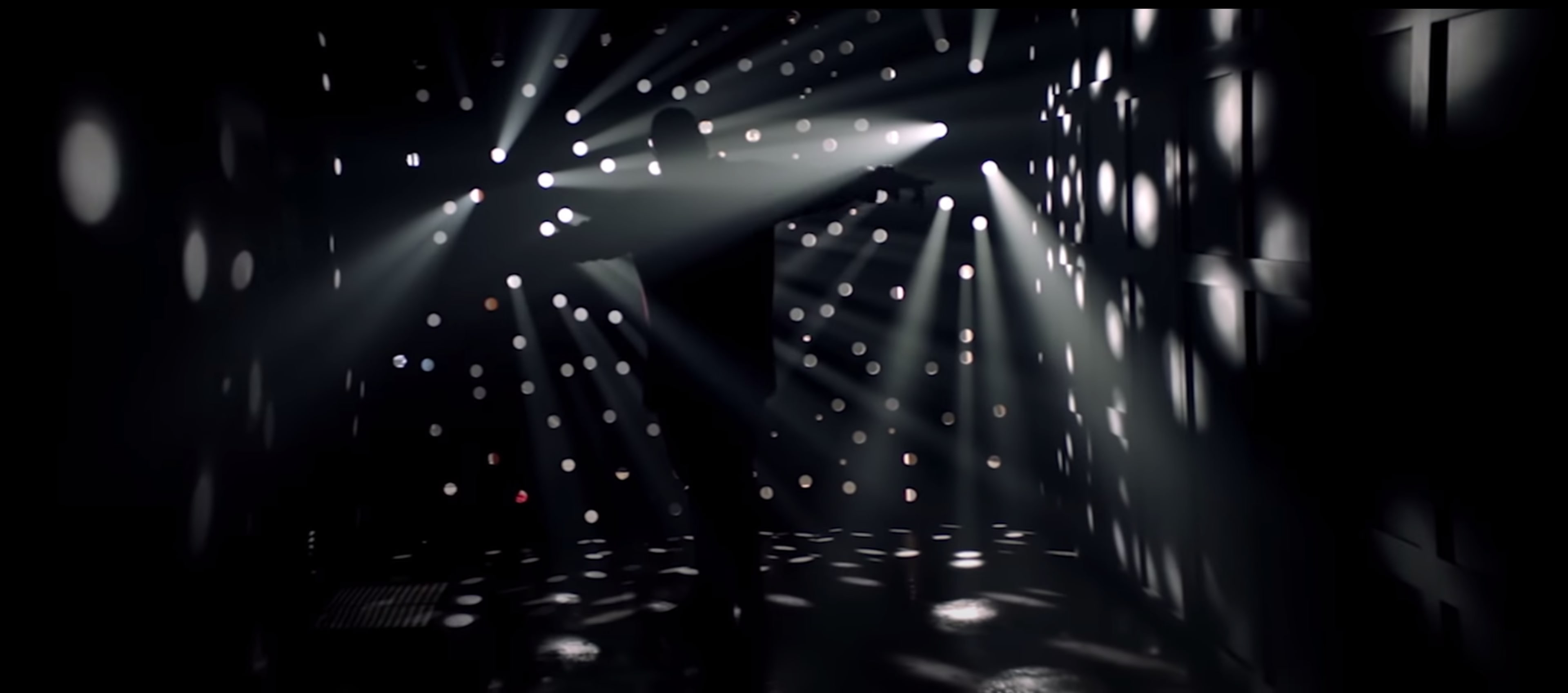 A figure stands in a dimly lit space, surrounded by beams of light creating patterns on the walls.