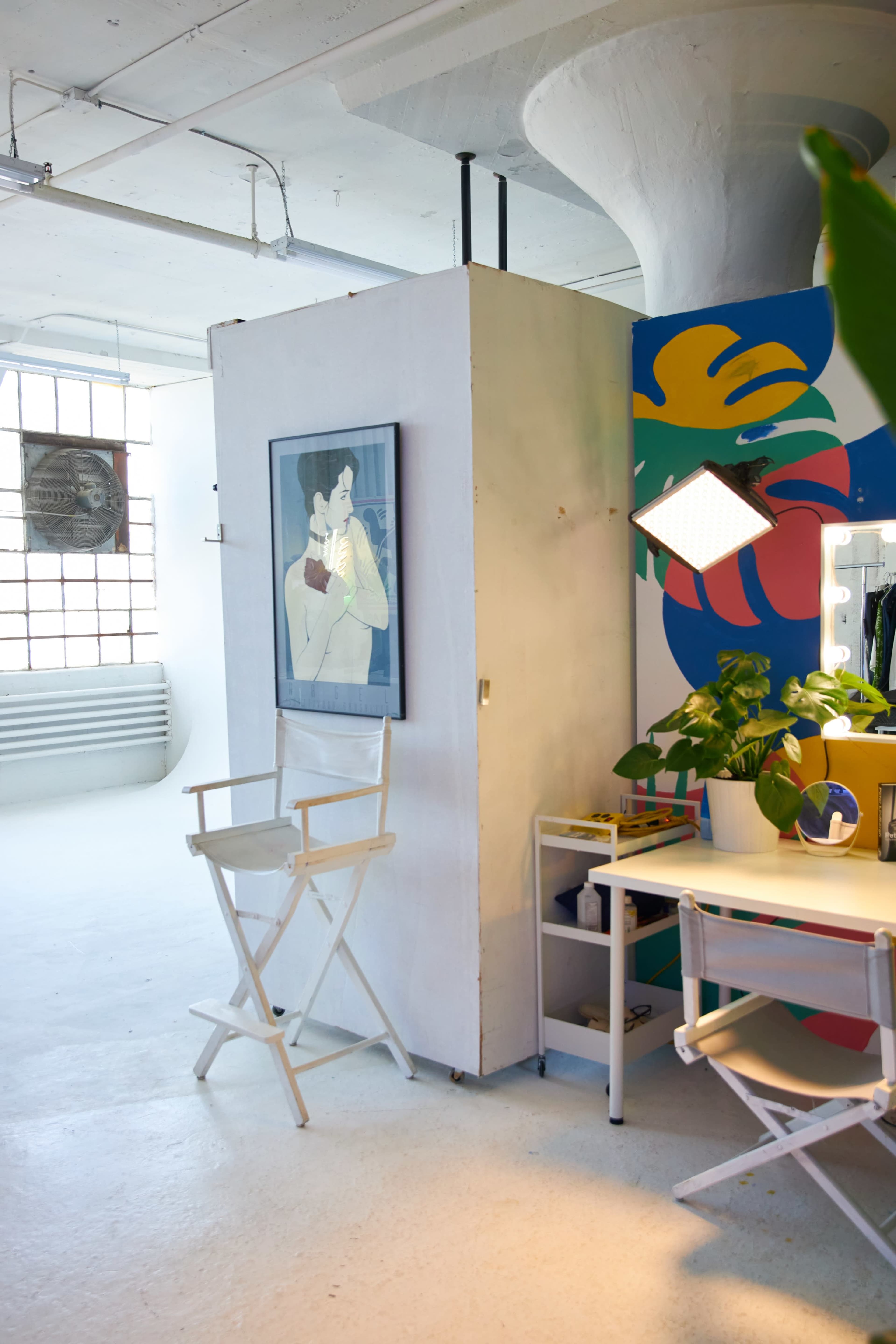 A bright, minimalist studio space featuring a painted wall, a modern chair, a small table with a mirror, and a large window letting in natural light.
