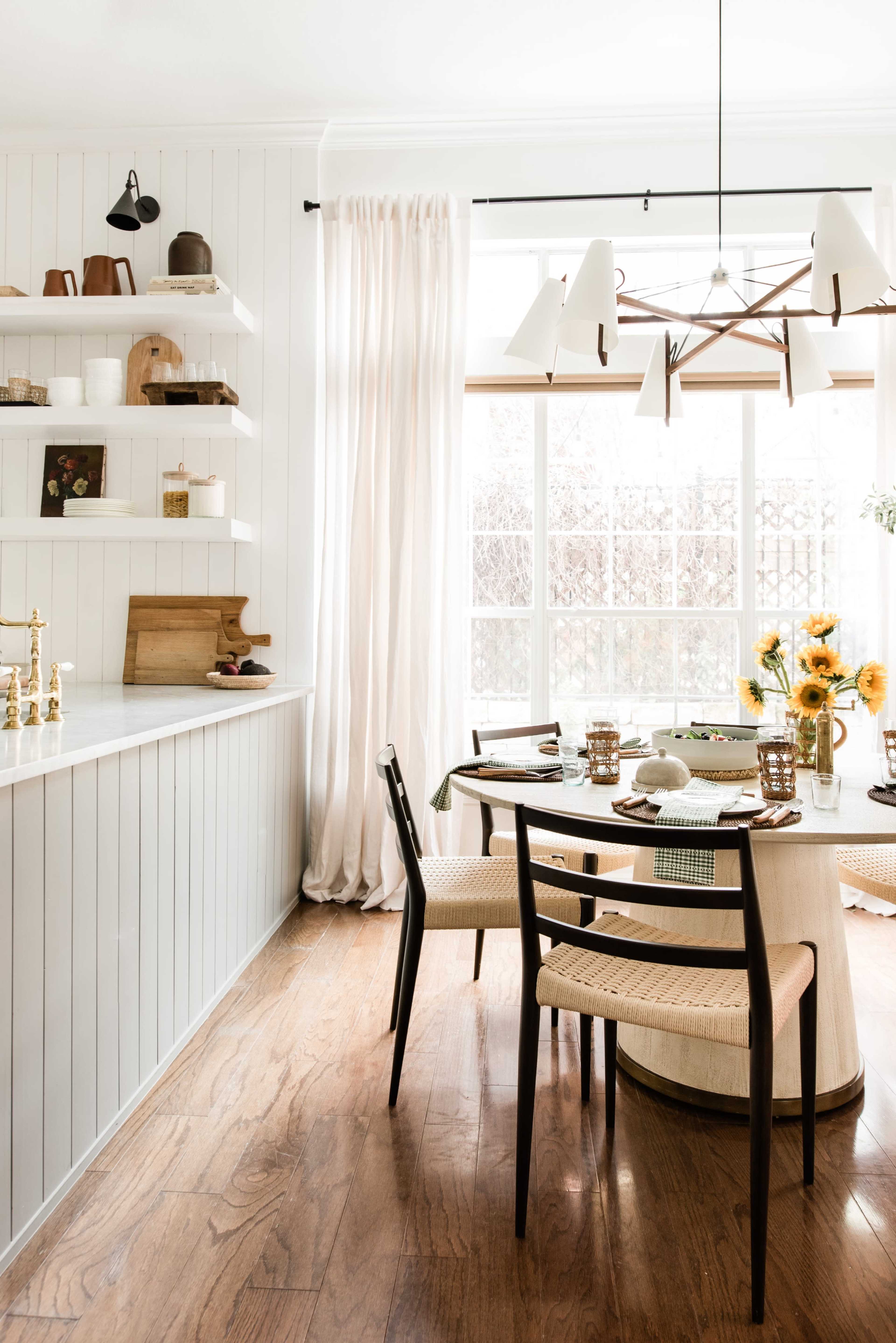 A dining area features a round table with woven chairs, a vase of sunflowers, and a shelf displaying various kitchen items by a large window with sheer curtains.
