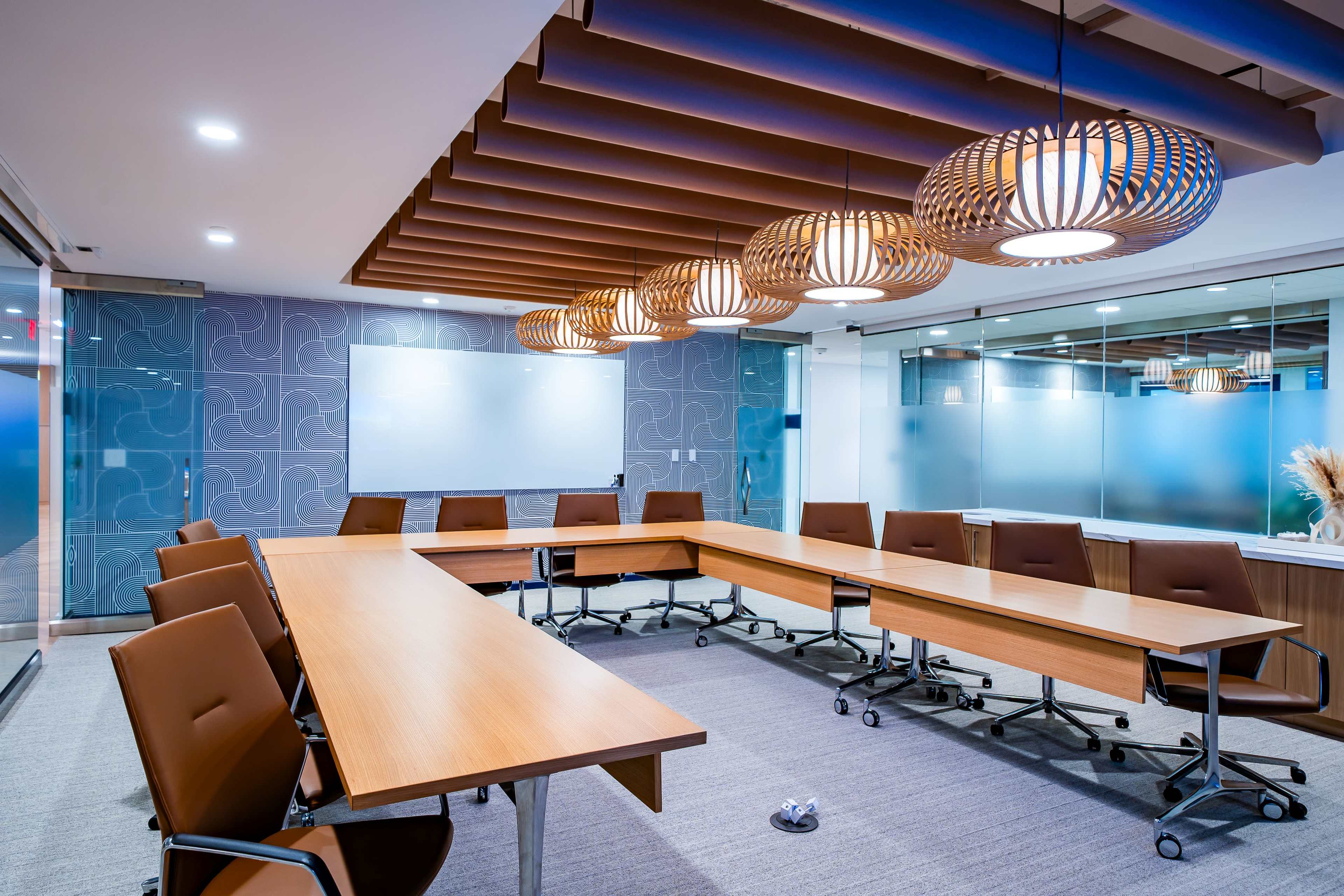 A modern conference room features a rectangular table surrounded by ergonomic brown chairs, with decorative pendant lights hanging from a wooden ceiling.