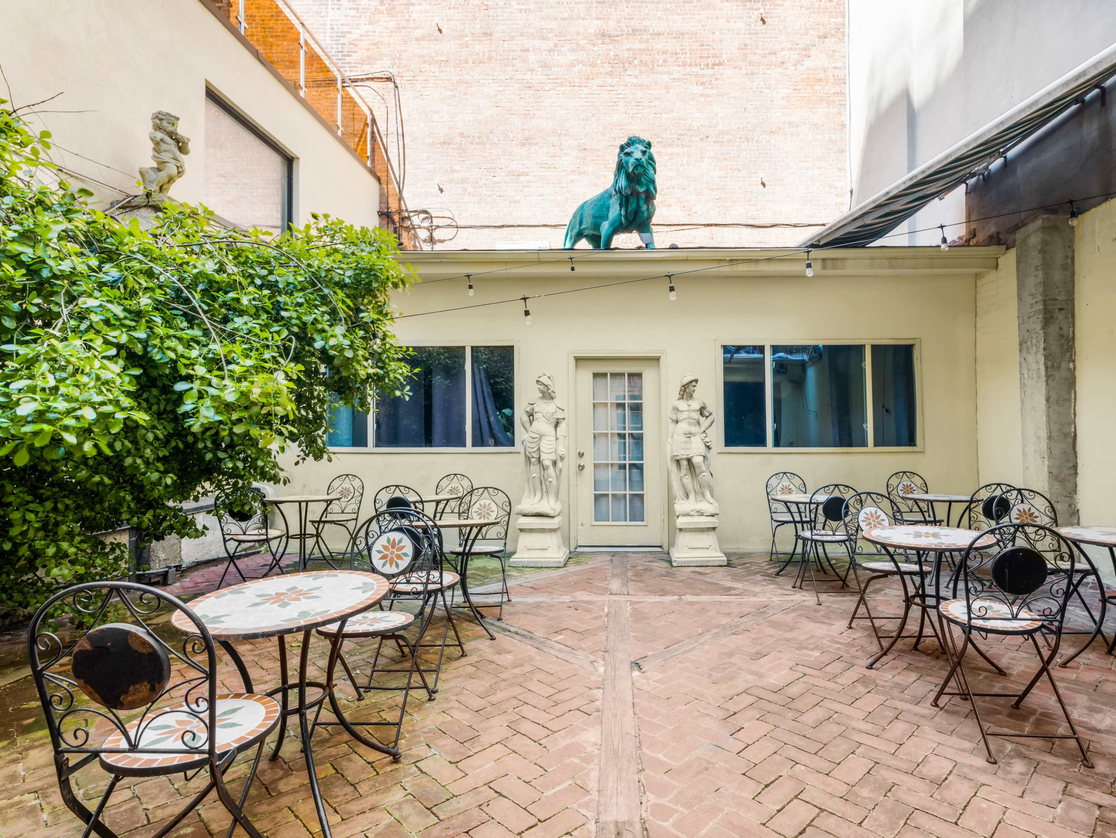 The image shows a small outdoor courtyard featuring several round tables with wrought iron chairs, flanked by two statues and a green lion sculpture on the roof.