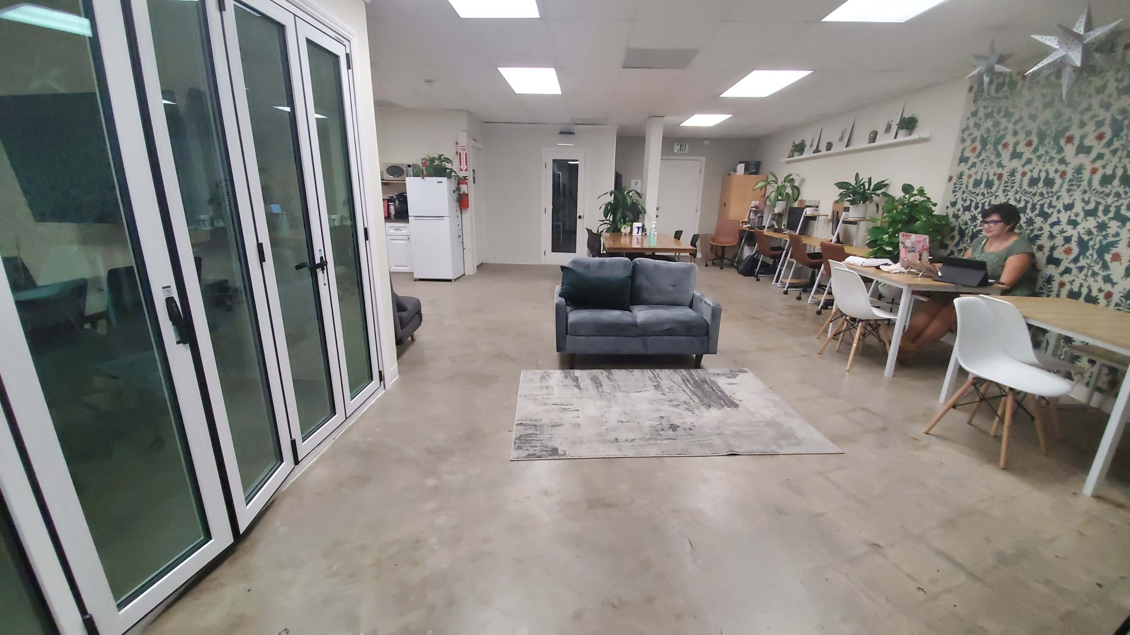 The image shows a modern office space with a gray sofa, a rug, a mix of tables and chairs, and large glass doors leading to an area with plants and a kitchenette.