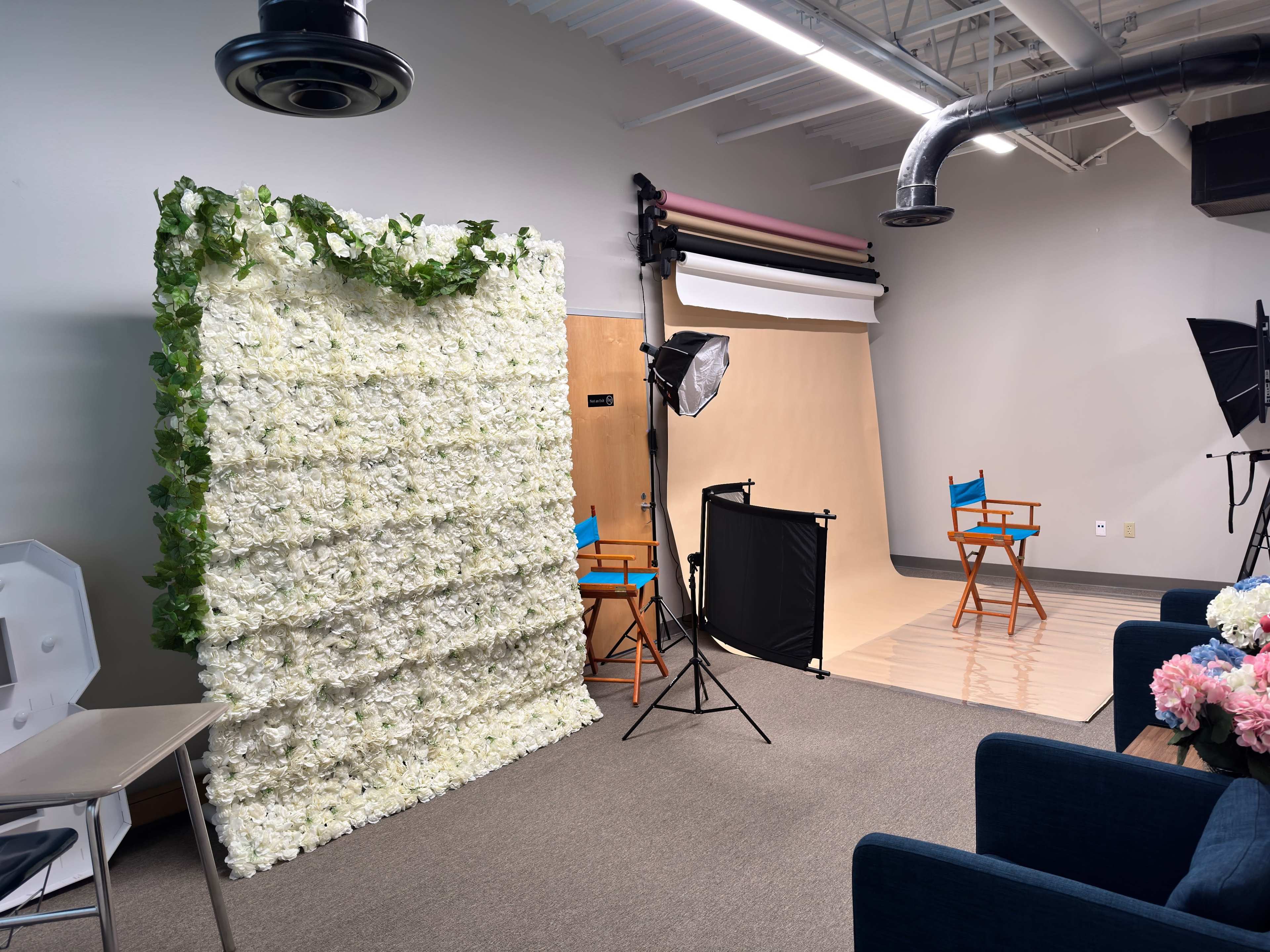 The image shows a photography studio setup featuring a flower wall backdrop, two director's chairs, and various photography equipment.
