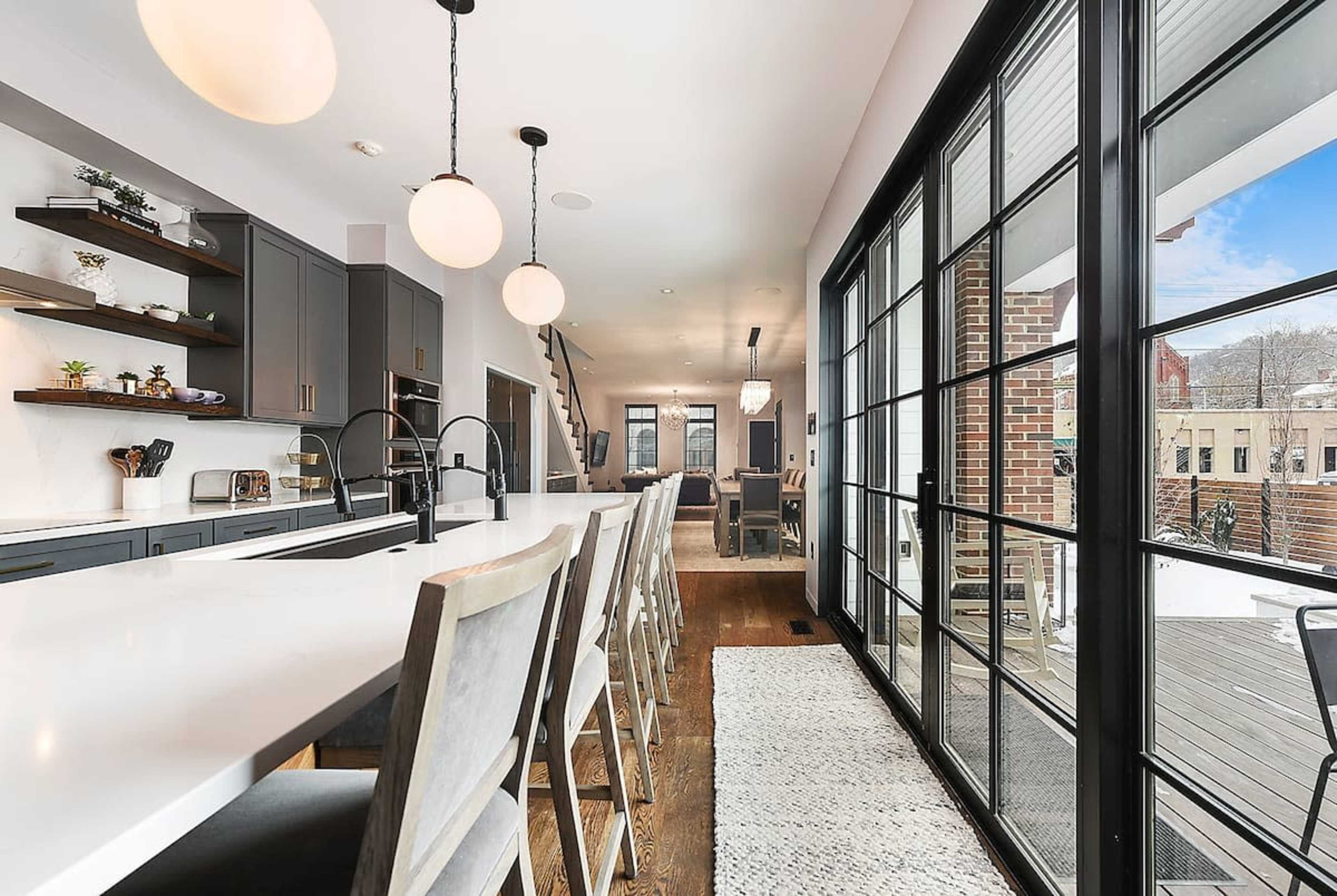 A modern kitchen with a long white countertop, gray cabinetry, and large windows leading to an outdoor space.